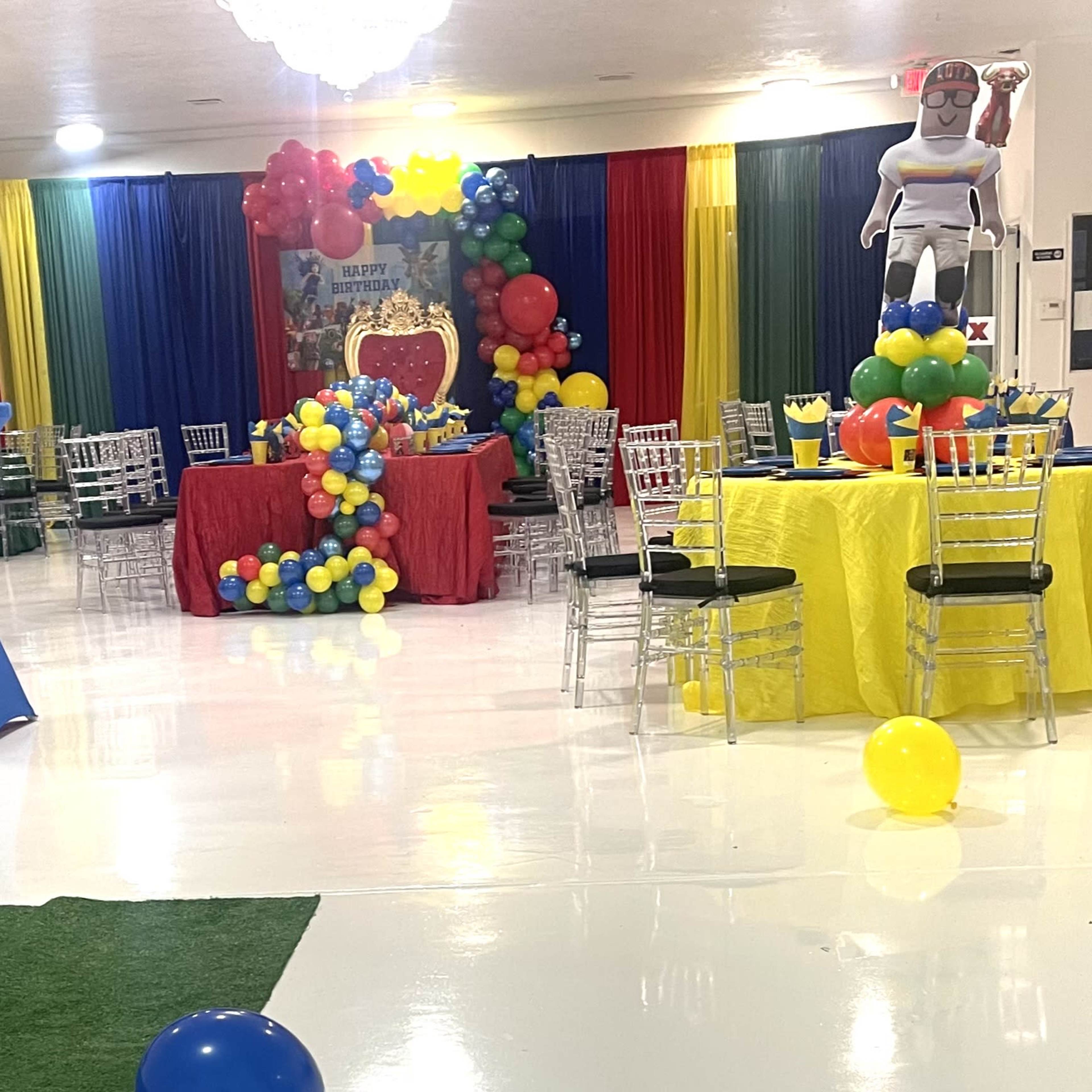 A colorful banquet hall is set up with decorated tables and chairs, featuring balloons and a large centerpiece in a festive arrangement.