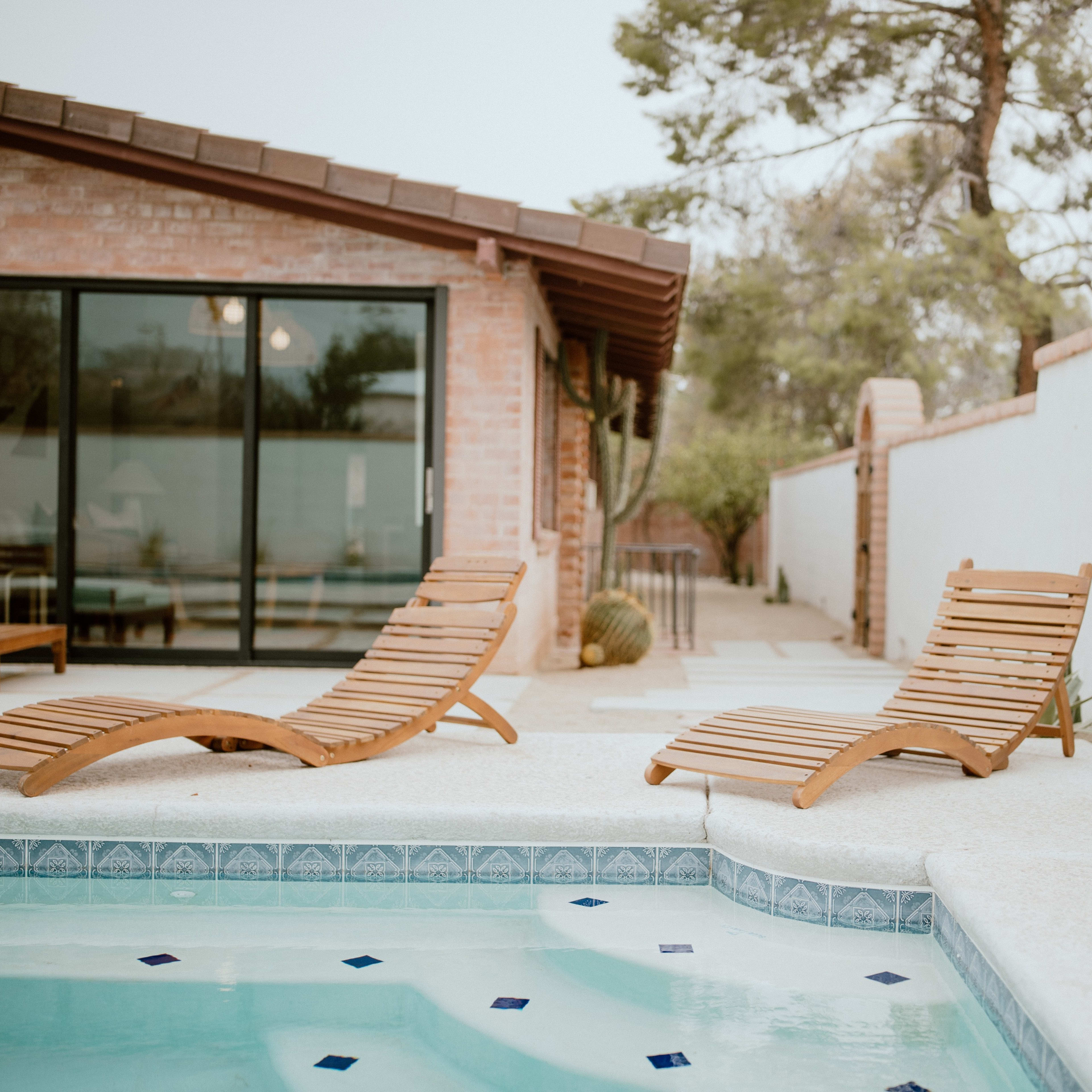 Two wooden lounge chairs rest by a swimming pool in a landscaped backyard, with a wall and cacti in the background.