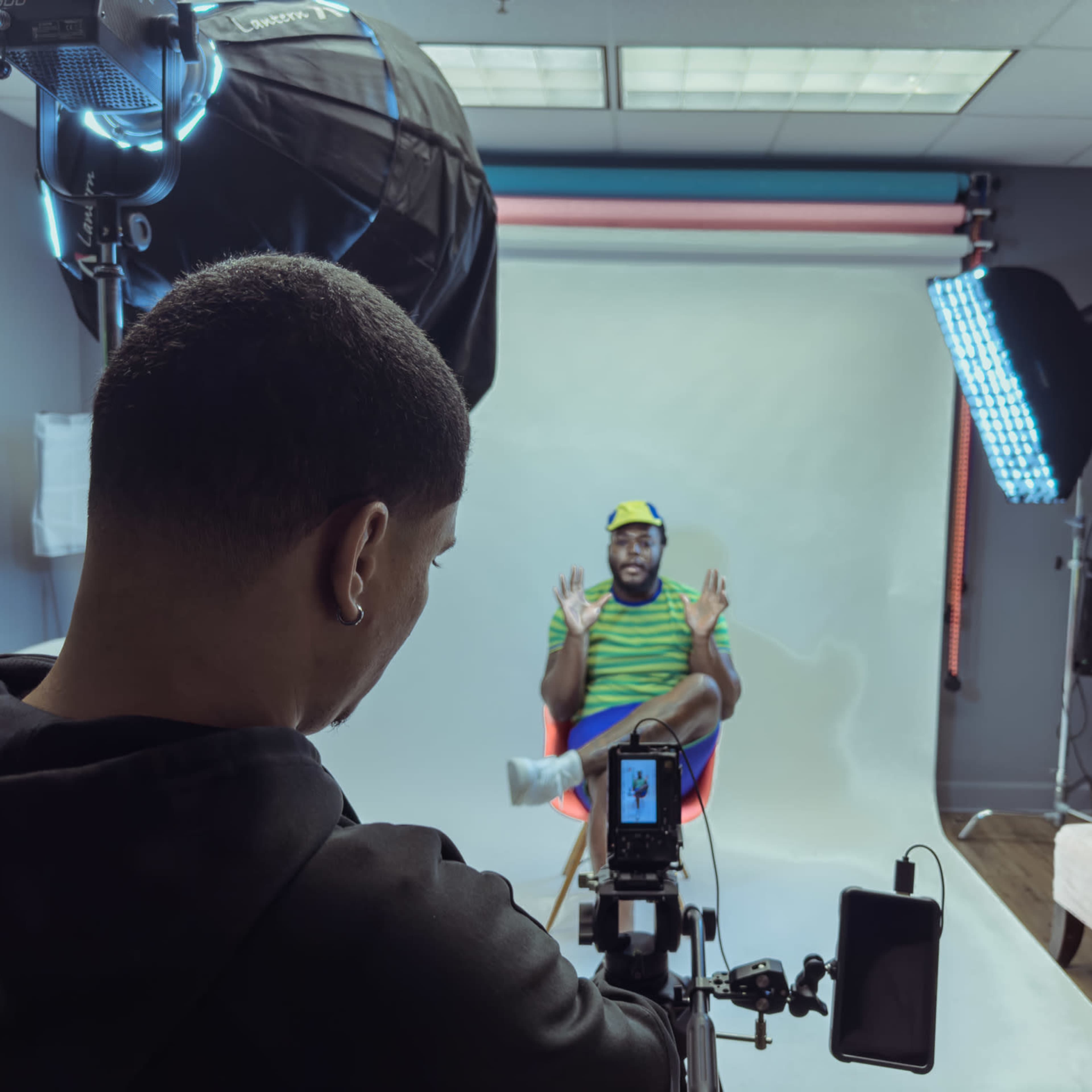 A person is filming an interview in a studio setting with a subject sitting in a chair against a backdrop.