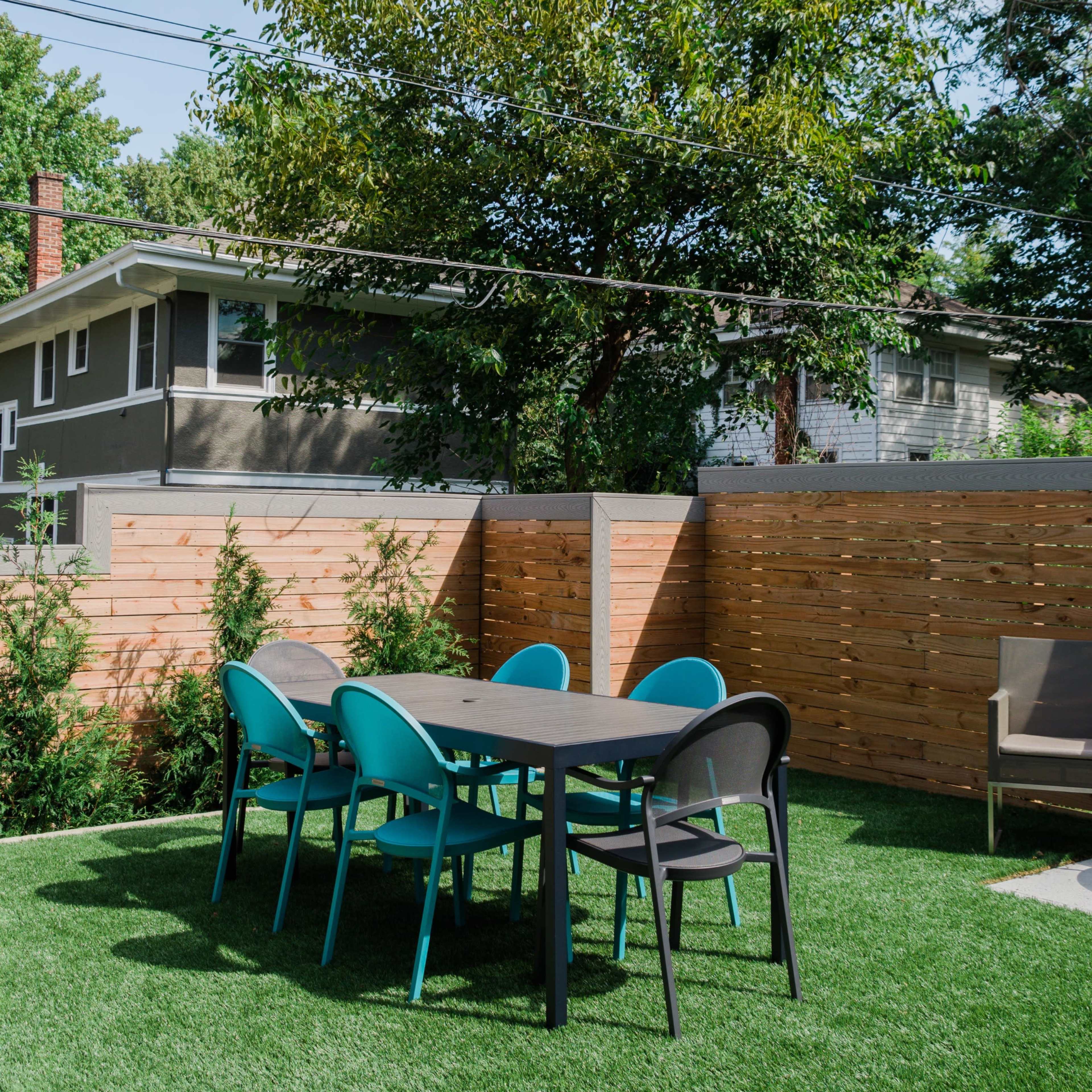 A backyard features a set of teal and black outdoor chairs gathered around a rectangular table on green synthetic grass, surrounded by wooden fencing and trees.