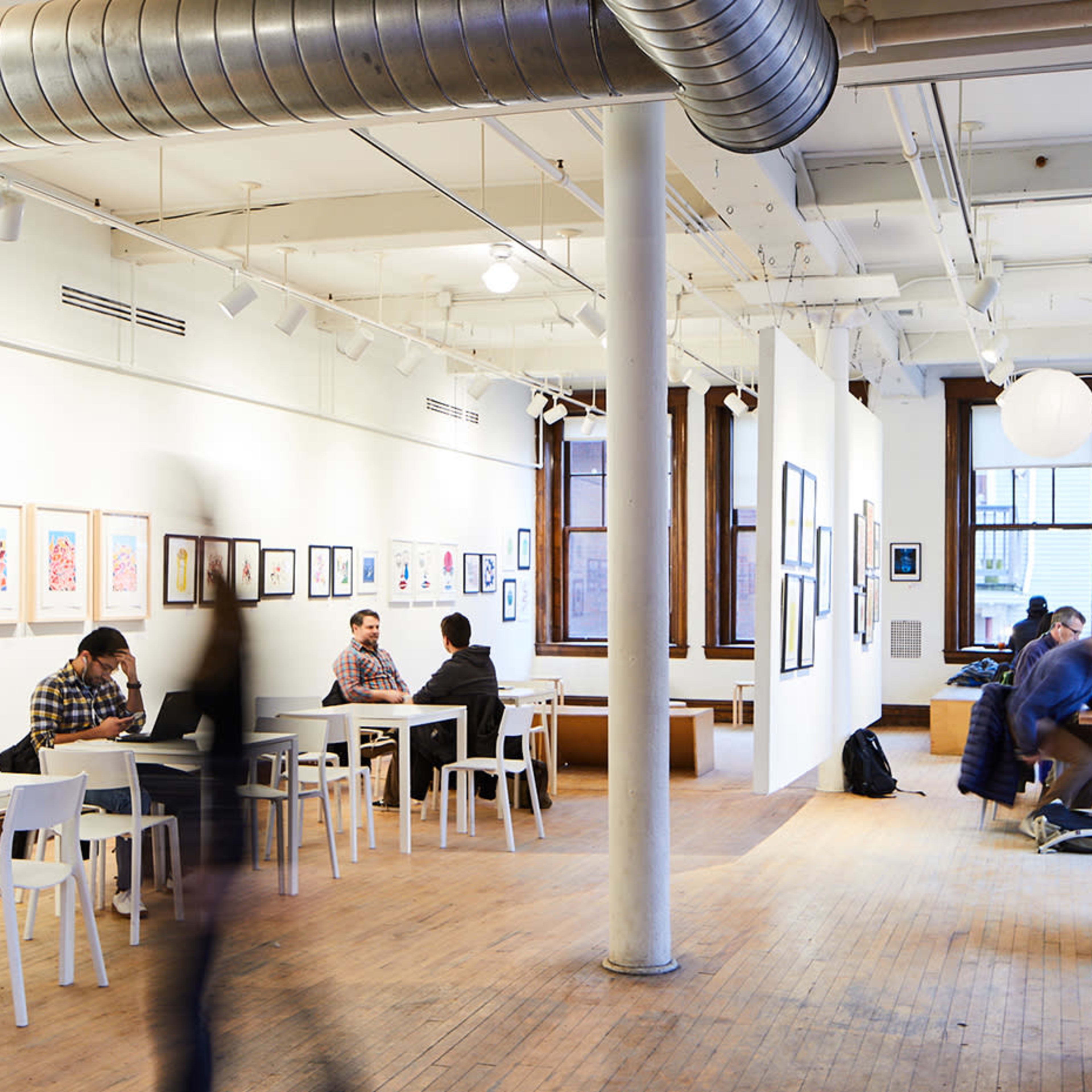 An art studio featuring tables occupied by people, with artwork displayed on the walls and large paper lanterns hanging from the ceiling.