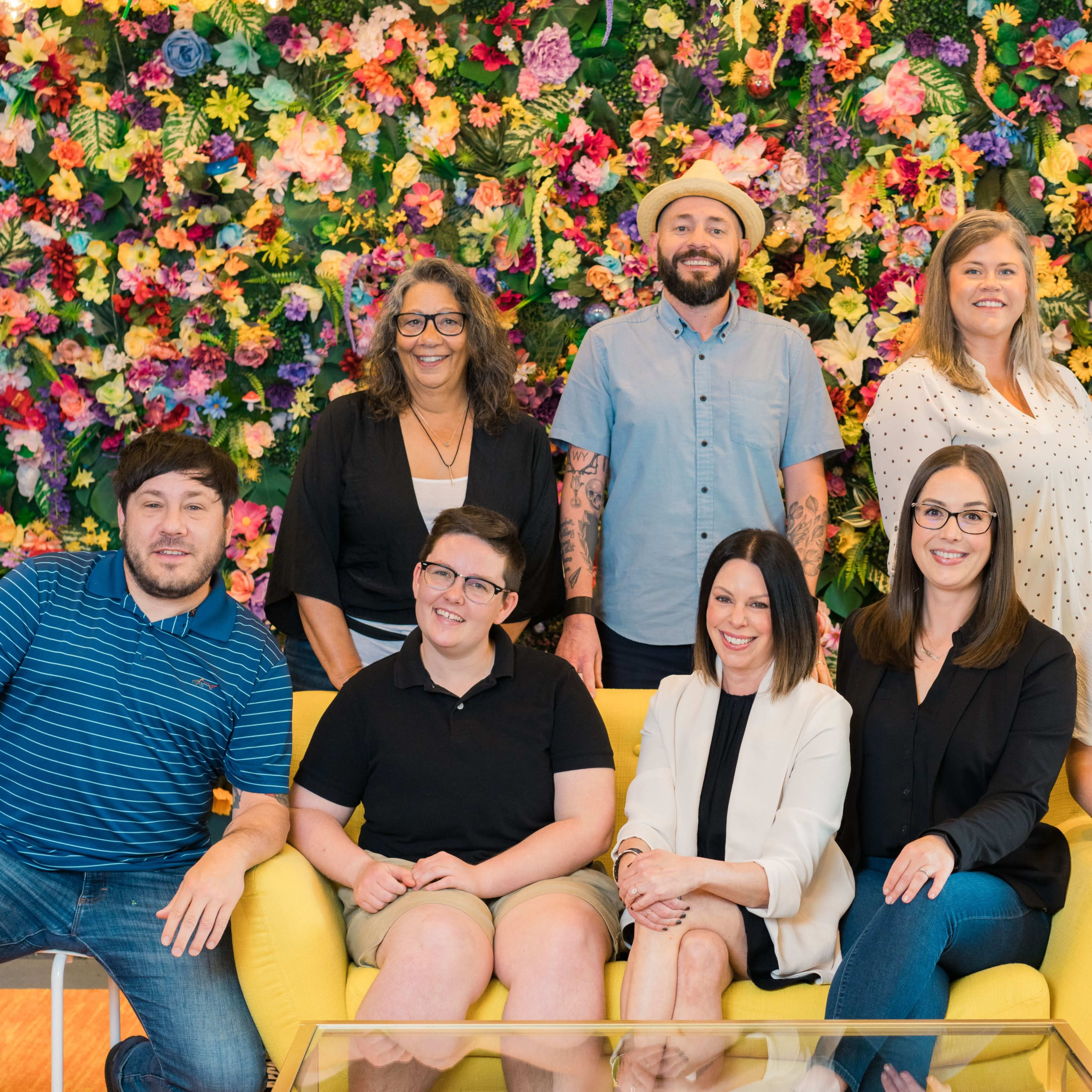 A group of eight individuals poses together in front of a vibrant floral wall, seated on a yellow sofa.