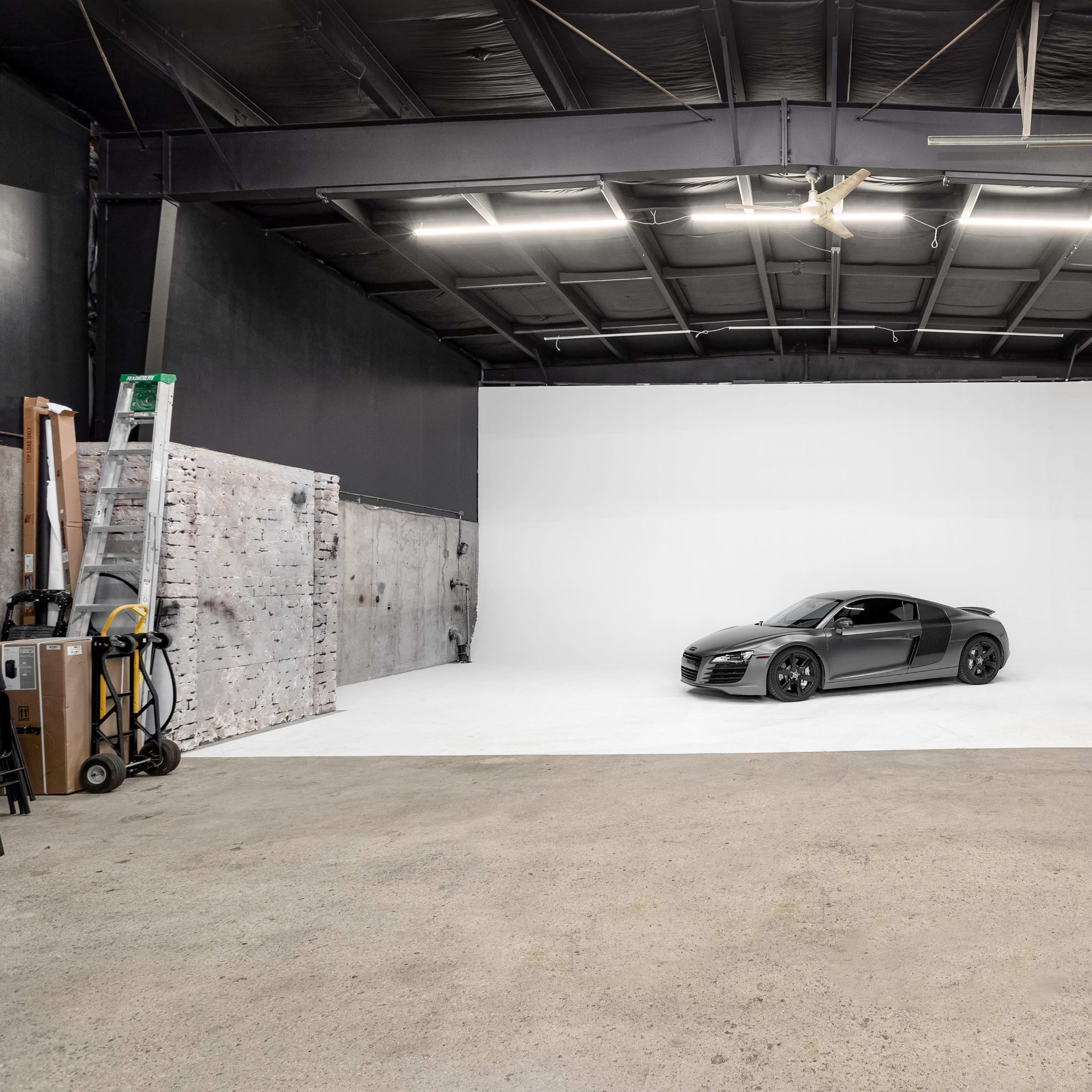 A sleek gray sports car is positioned on a white backdrop inside a spacious studio with a concrete floor.