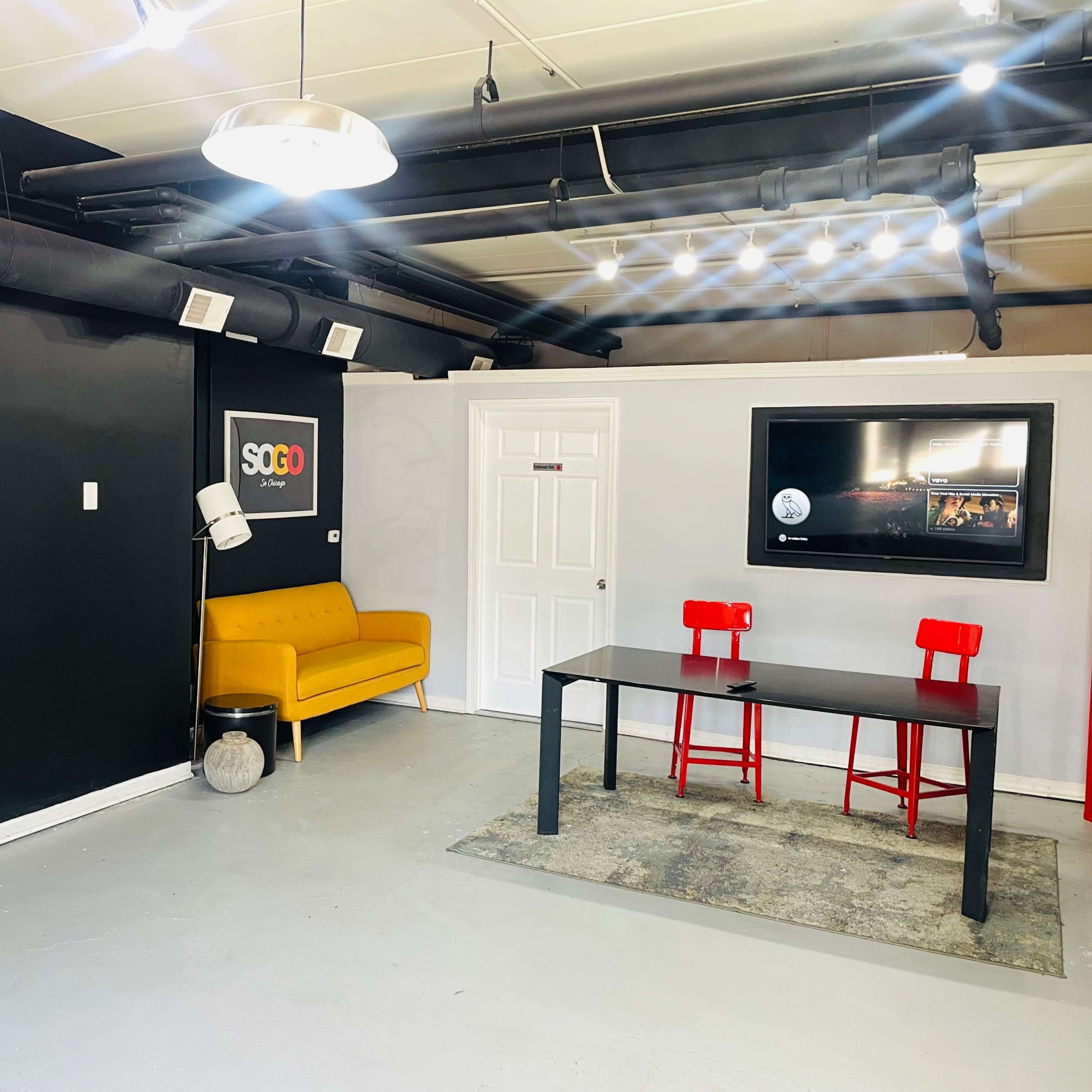 A modern indoor space featuring a yellow sofa, a black table with two red chairs, a wall with the words "SCHOOL HI. DID." in bold letters, and a television mounted on the wall.