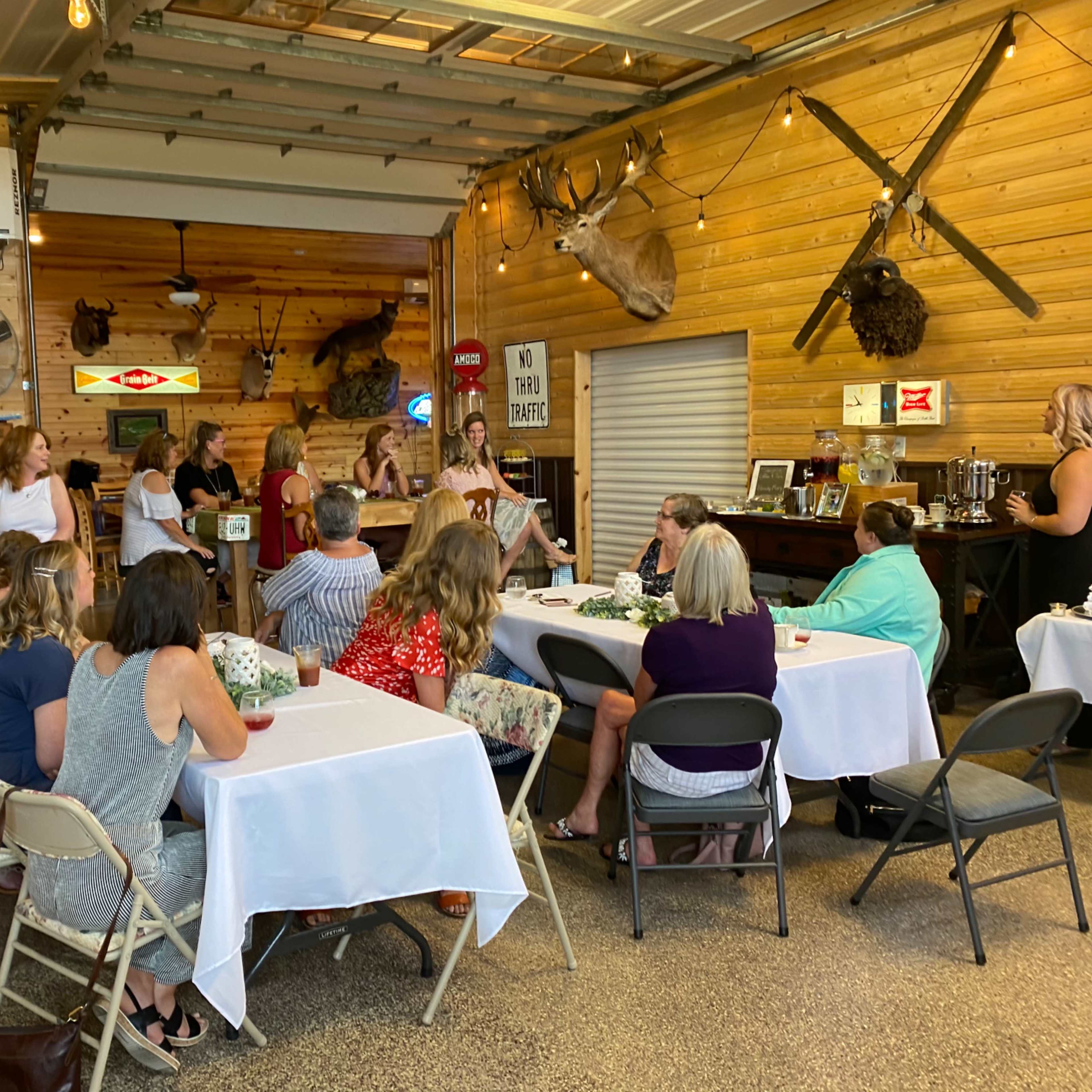 A group of individuals sits around tables in a wooden-paneled room decorated with mounted trophies and vintage signs.