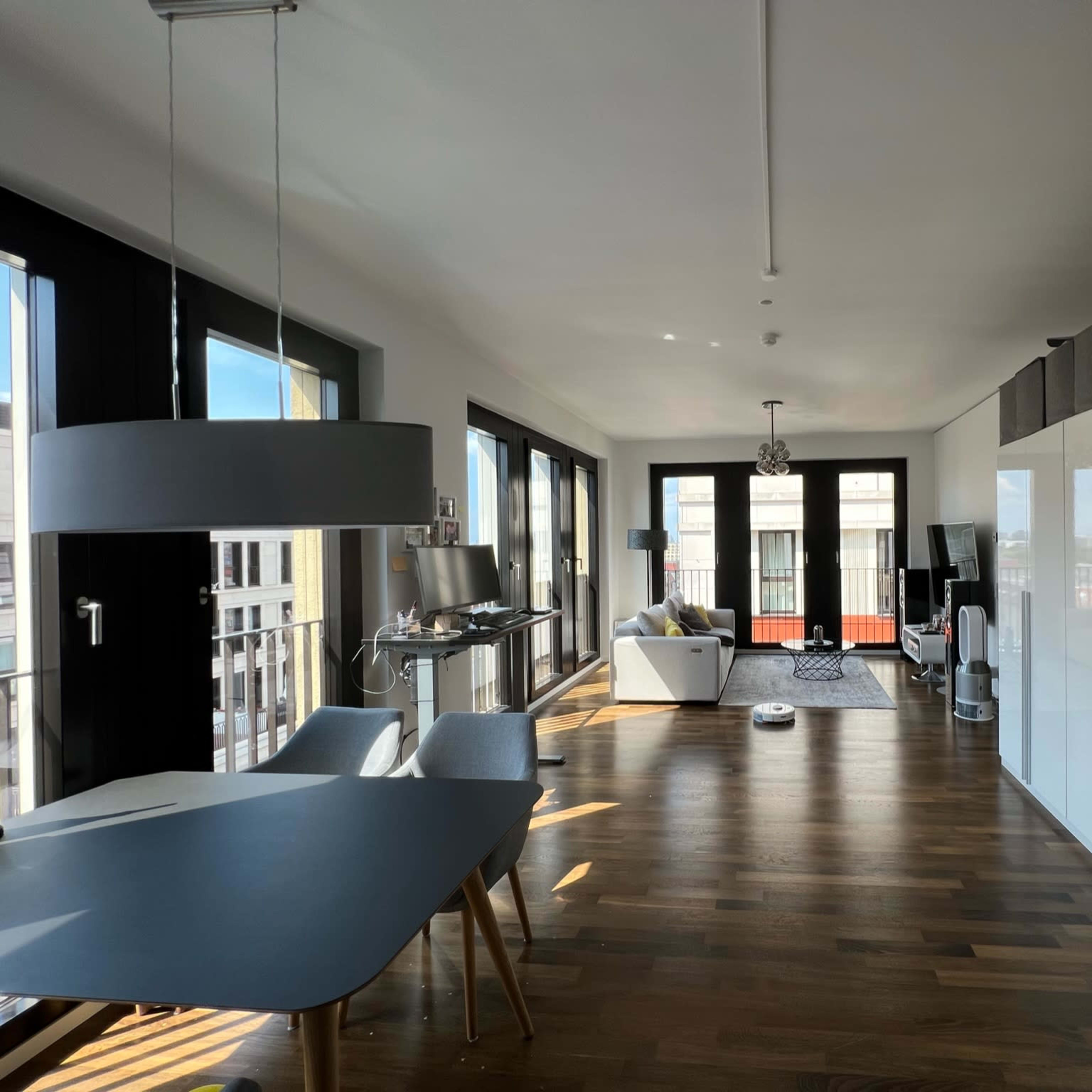 A modern, open living space with a dining area featuring a table and chairs, a white sofa, and large windows allowing natural light.