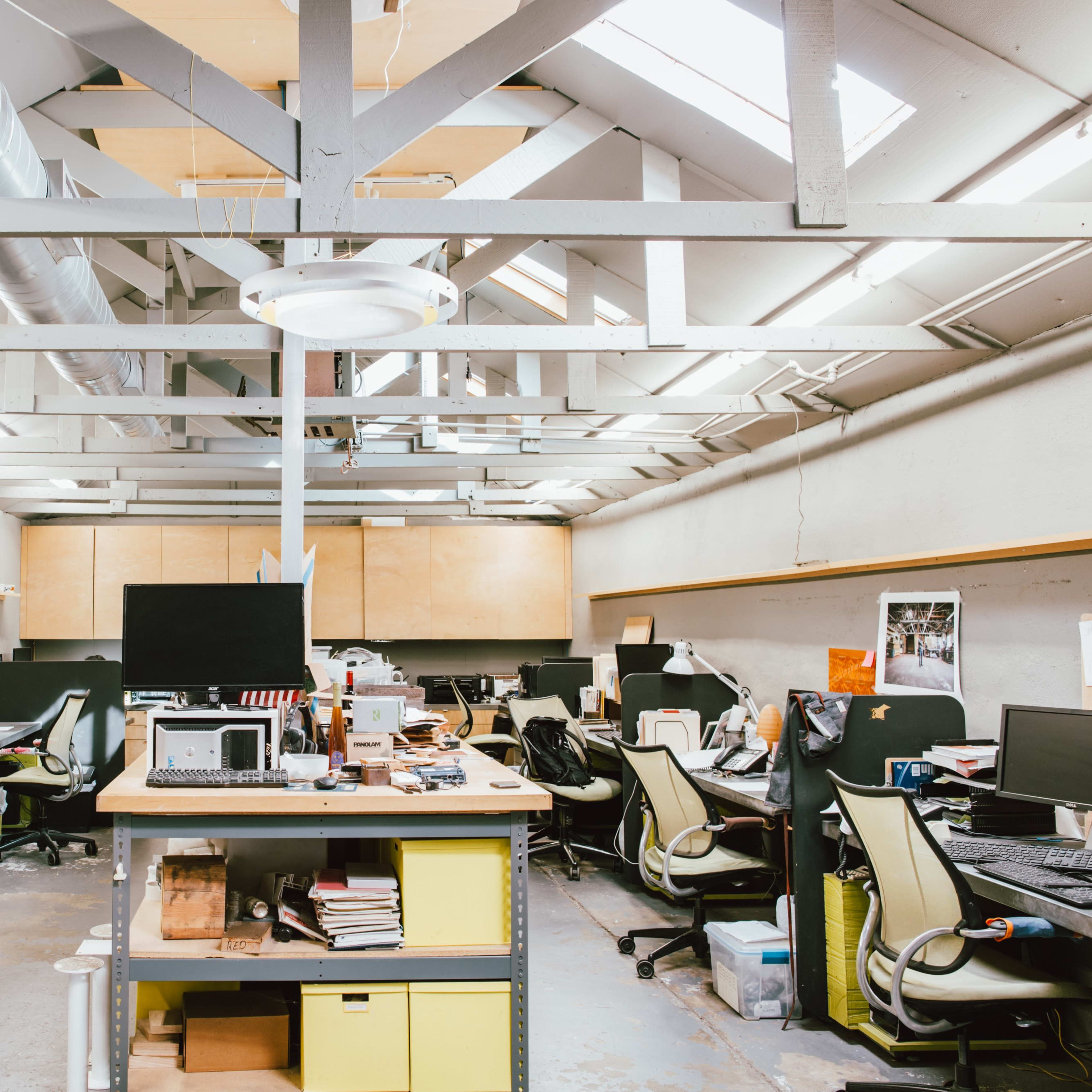 An open office space with multiple workstations, each equipped with computers and office supplies, under a vaulted ceiling with exposed beams.