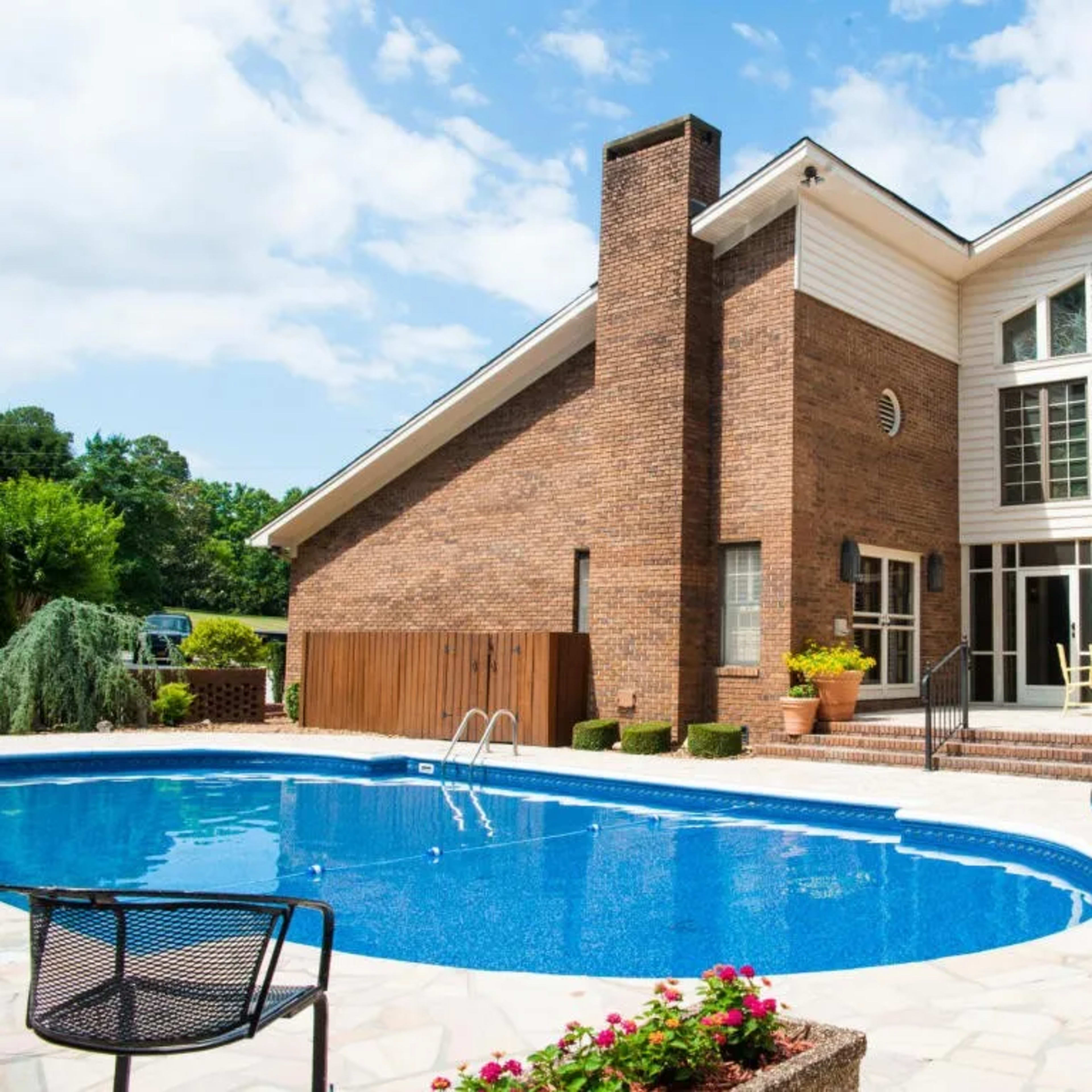 A brick house with large windows overlooks a blue swimming pool surrounded by stone patio and greenery.