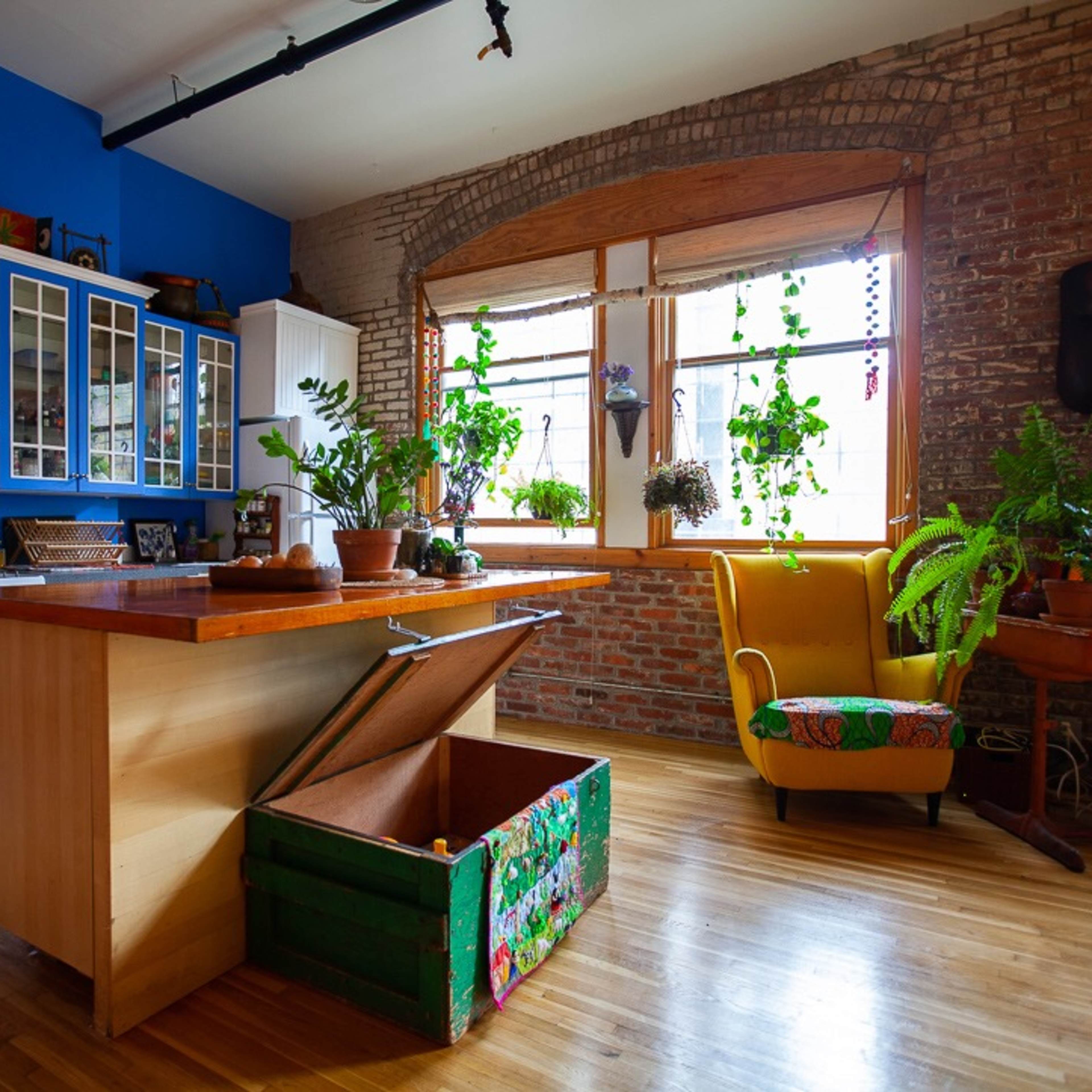 A cozy kitchen with a wooden island, a gas stove, and large windows adorned with various indoor plants, set against exposed brick walls.