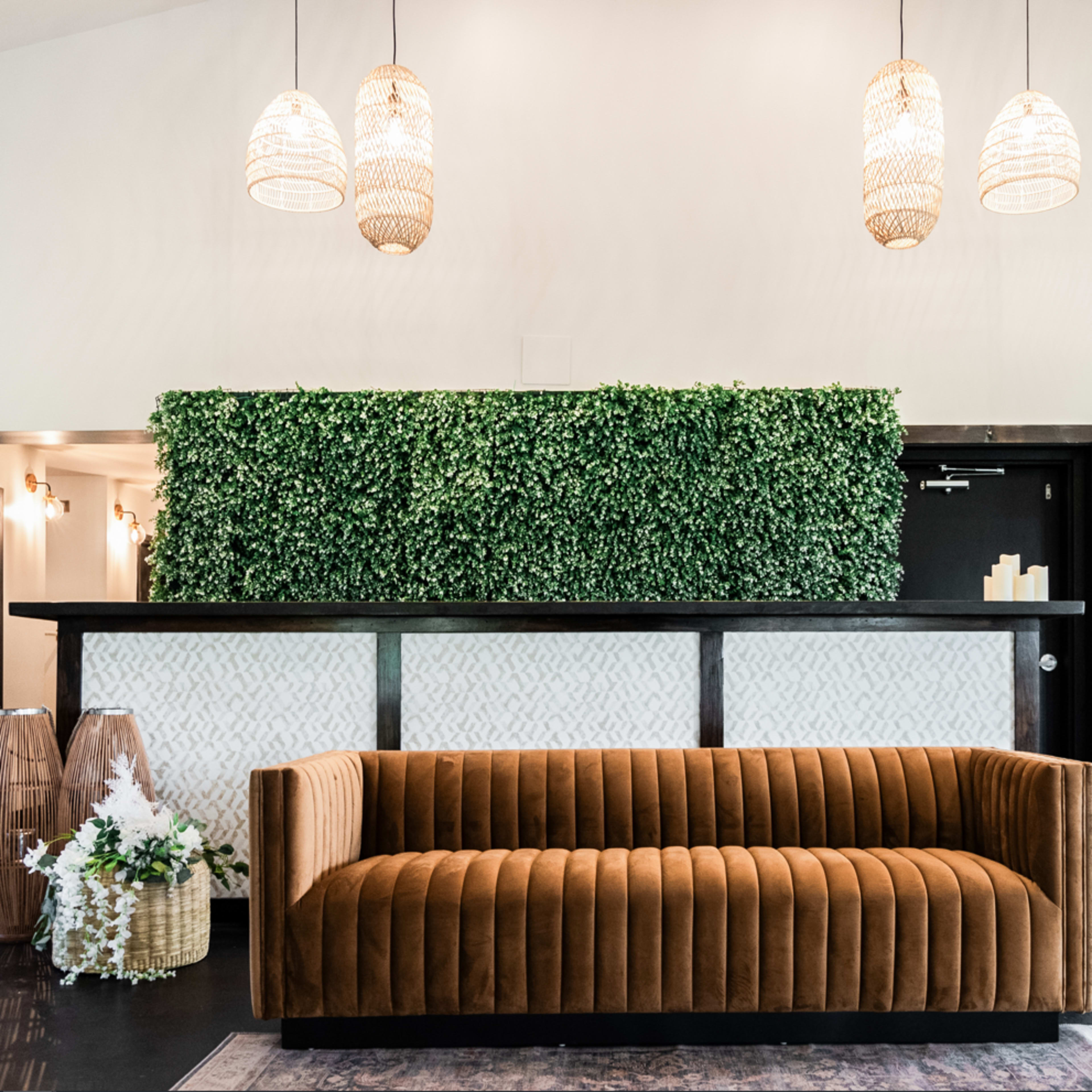 A modern reception area featuring a brown velvet sofa, a green leafy panel behind a wooden counter, and pendant lighting.
