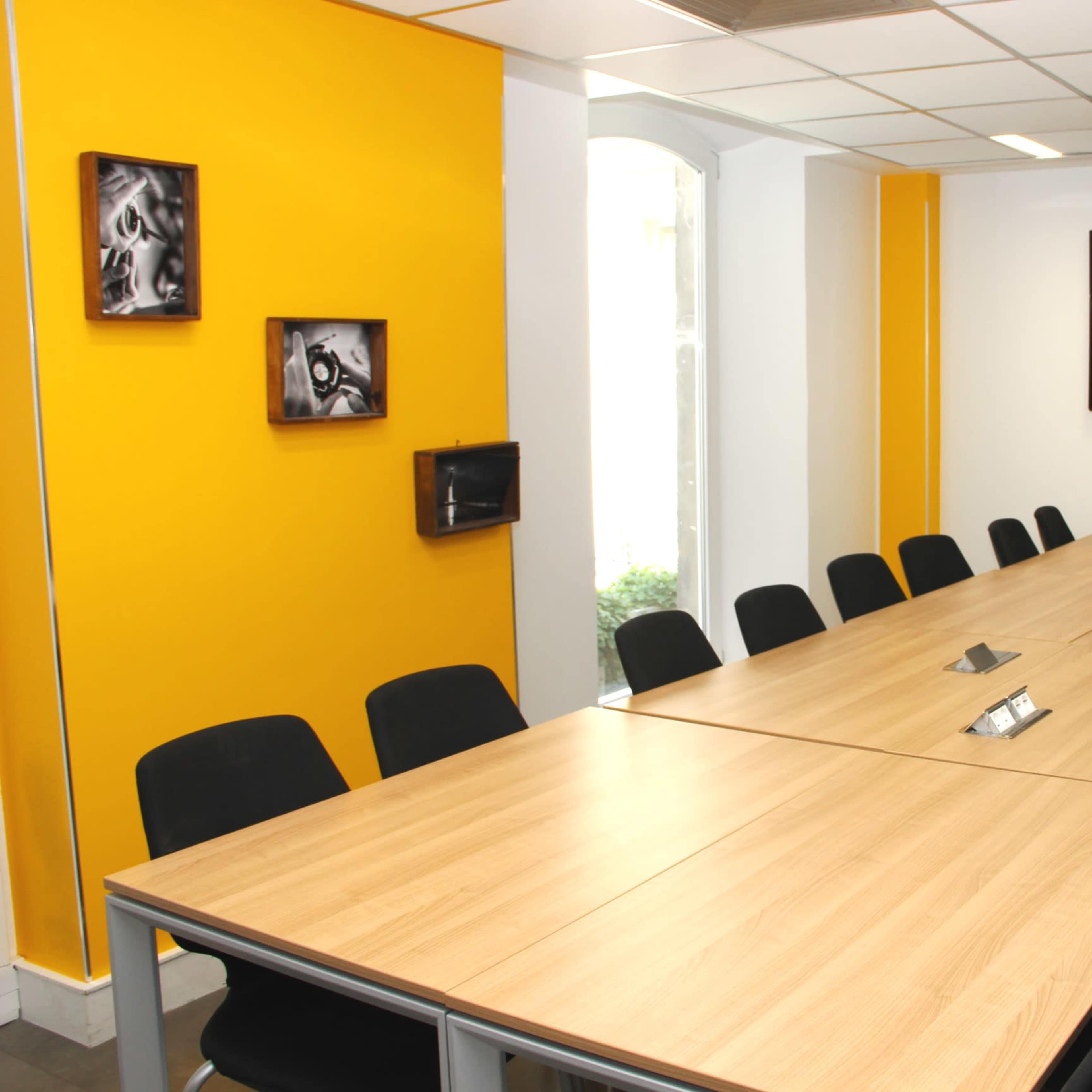 A meeting room with a long wooden table surrounded by black chairs, yellow accent walls, and a wall-mounted television.