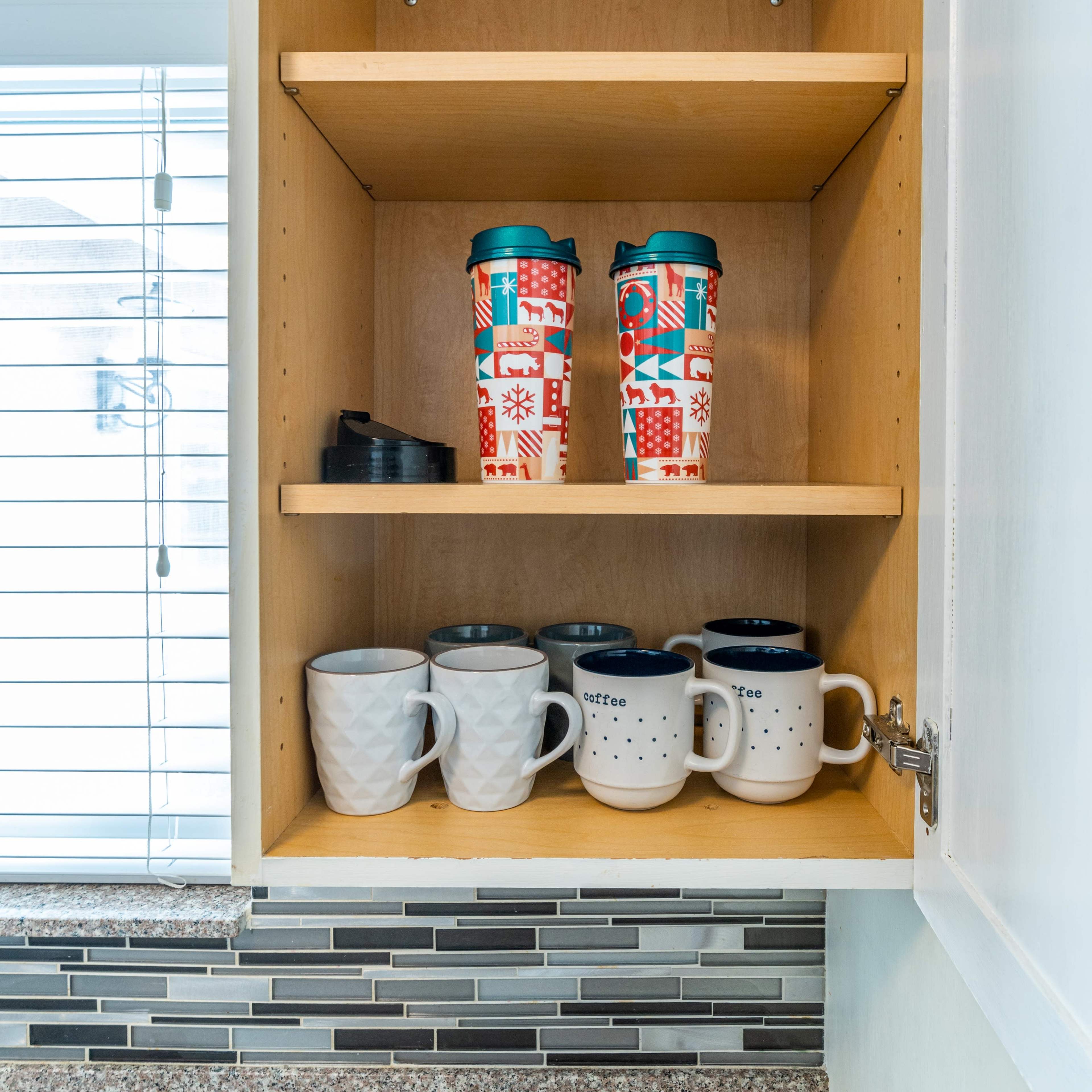 A kitchen cabinet contains mugs and travel tumblers organized on three shelves.