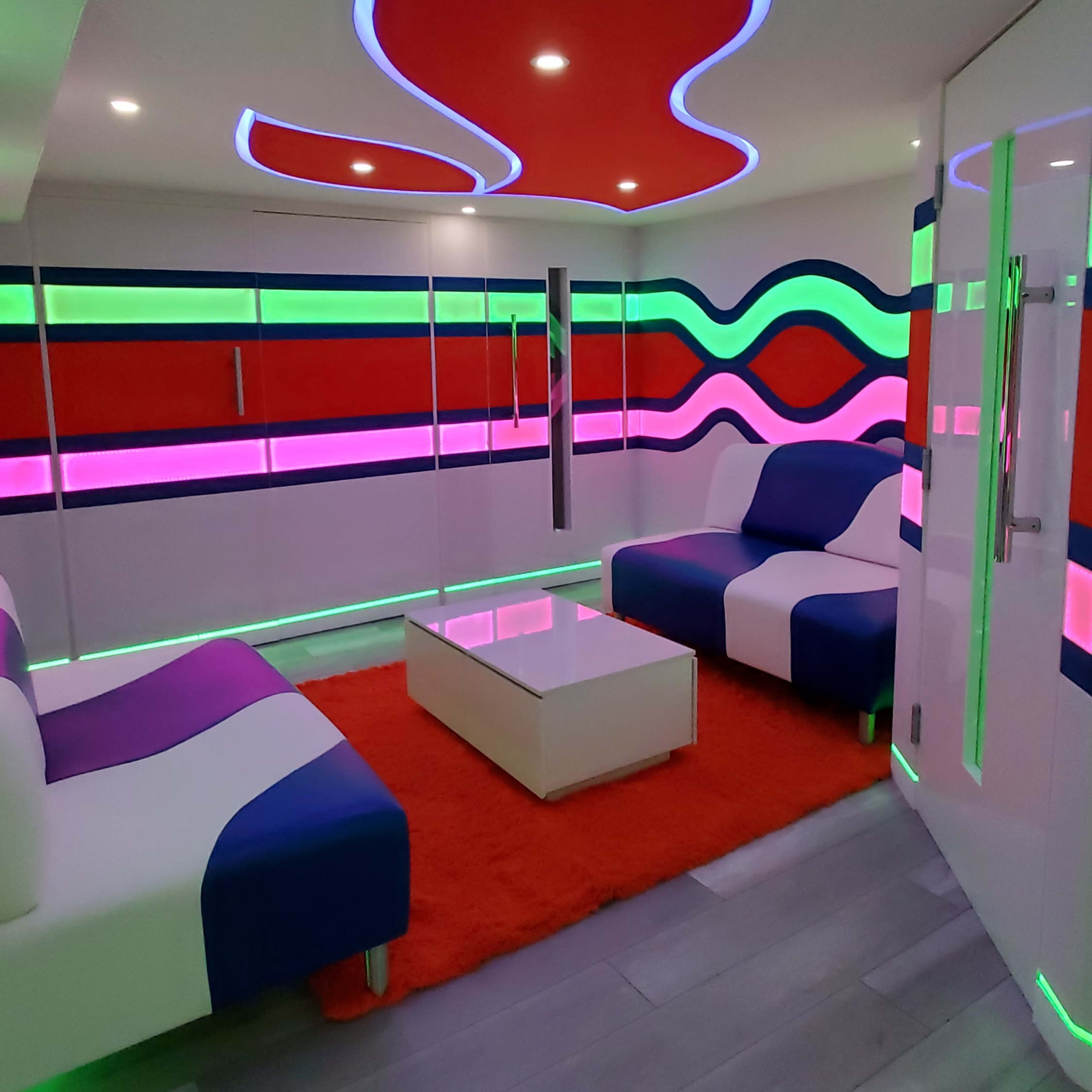 The room features bold, curving neon lights along the walls and ceiling, with modern furniture in a colorful design, including a white sofa with blue and purple accents on a vibrant red rug.