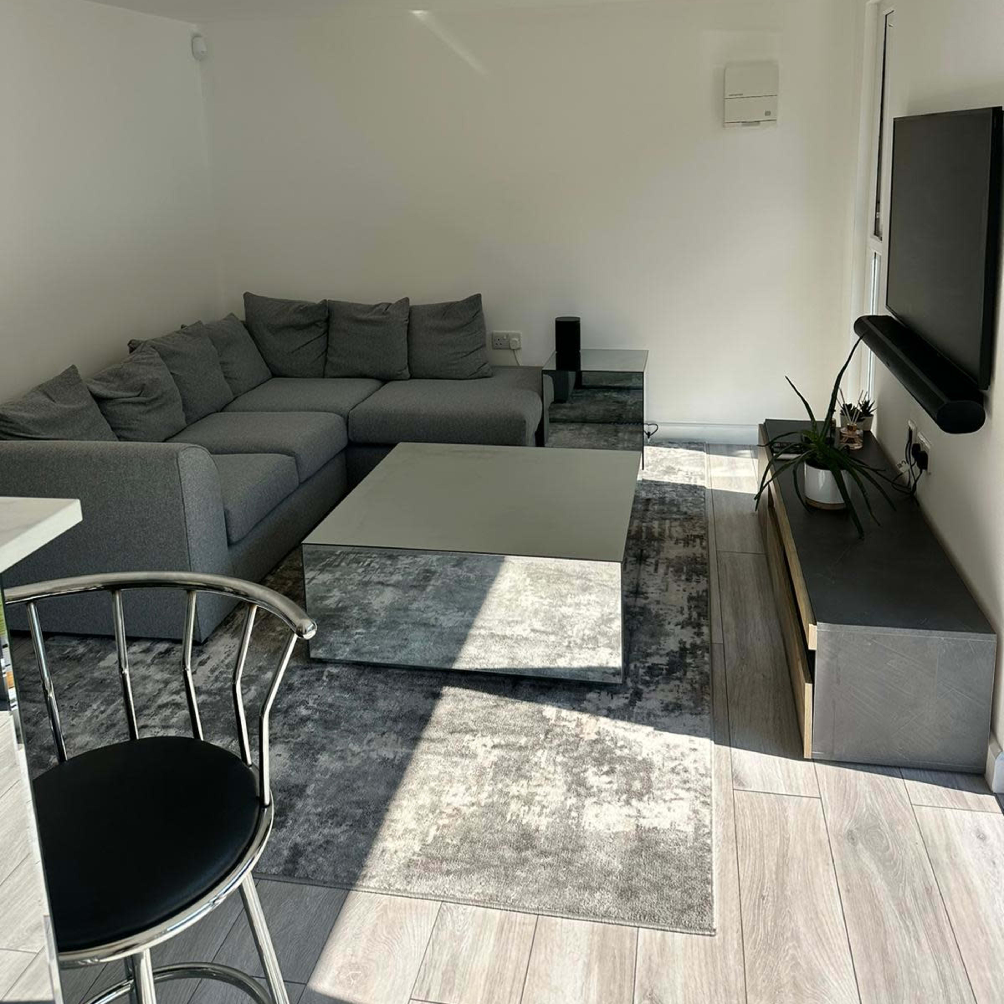 The scene shows a modern living room with a gray sectional sofa, a glass coffee table, a flat-screen TV mounted on the wall, and minimalistic decor.