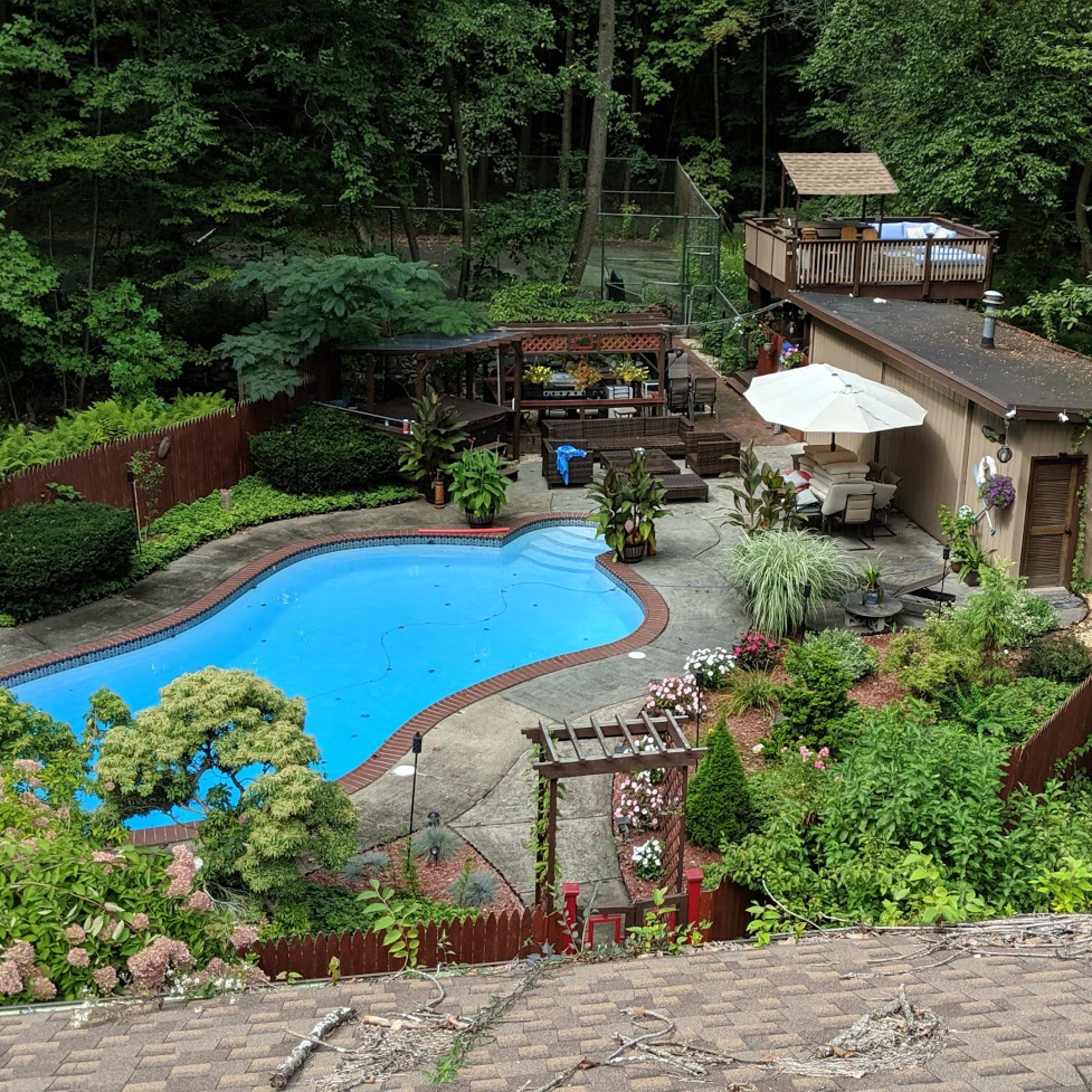 A landscaped backyard with a swimming pool, surrounded by lush greenery, wooden fencing, and various outdoor seating areas.