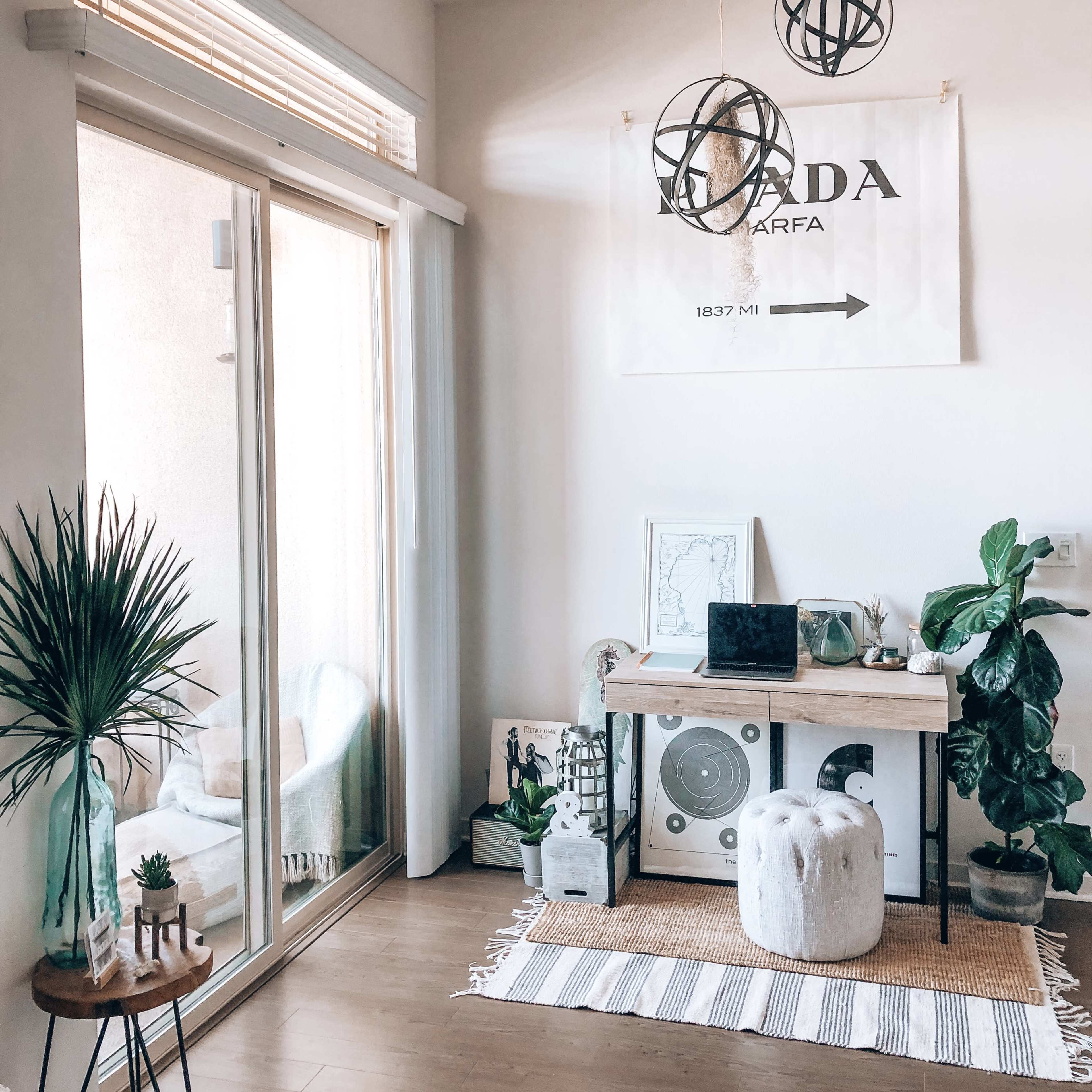 A bright and airy room featuring a workspace with a laptop on a table, surrounded by plants and decorative elements, along with a large window that allows natural light to enter.