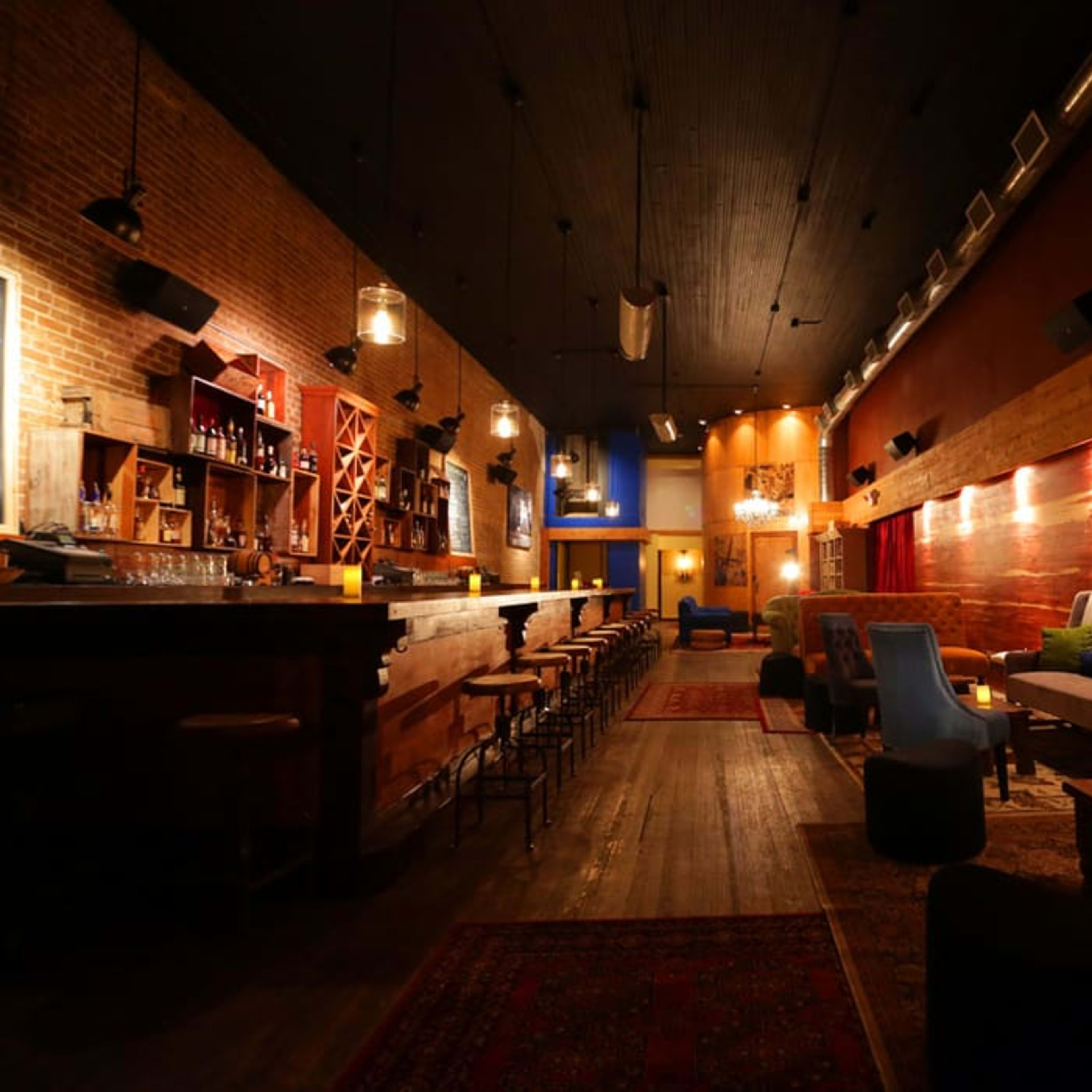 A dimly lit bar with a wooden interior, featuring a long bar counter, seating areas with plush chairs, and decorative lighting.