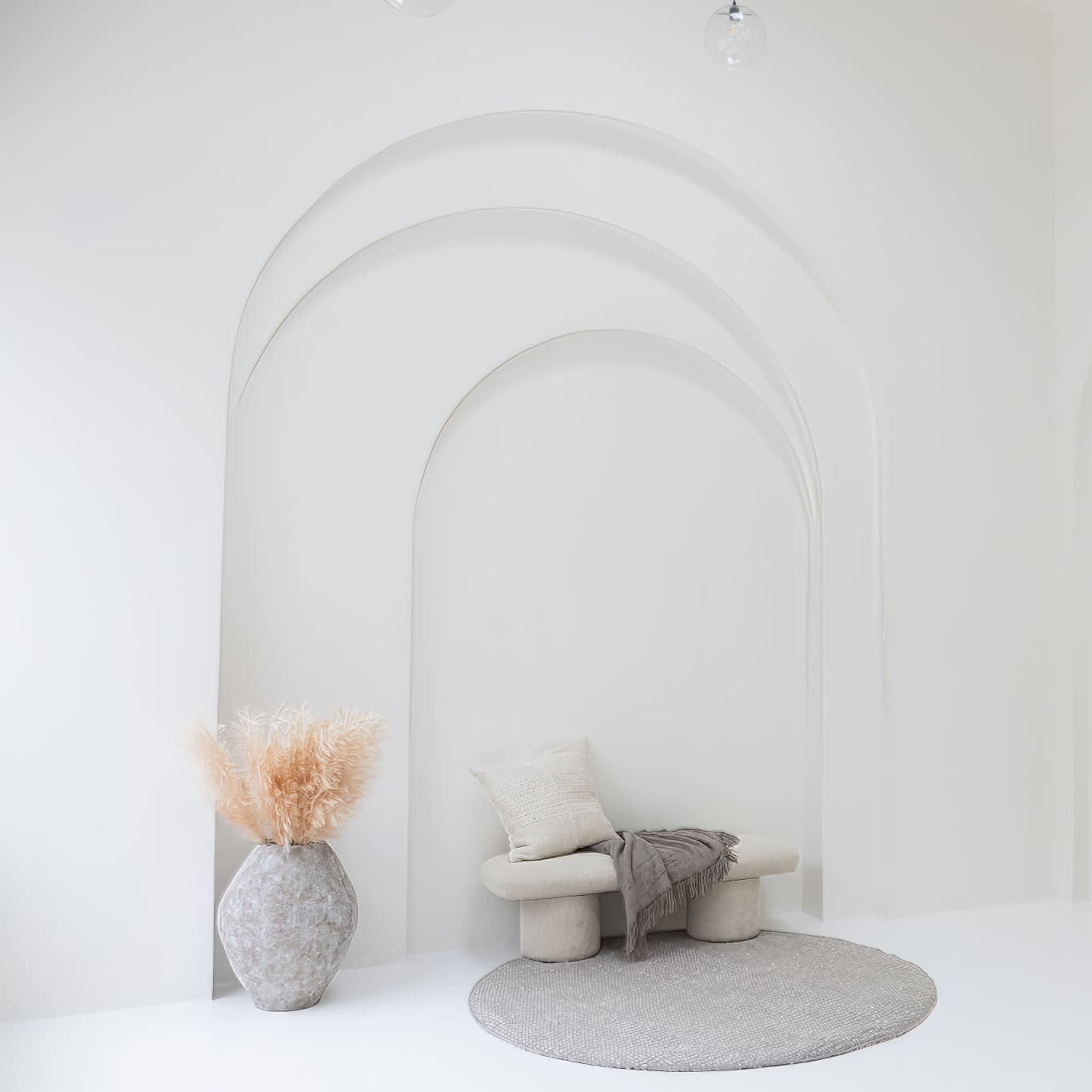 A minimalist interior with archways, a soft rug, a sculptural seating arrangement, and a vase with dried foliage.