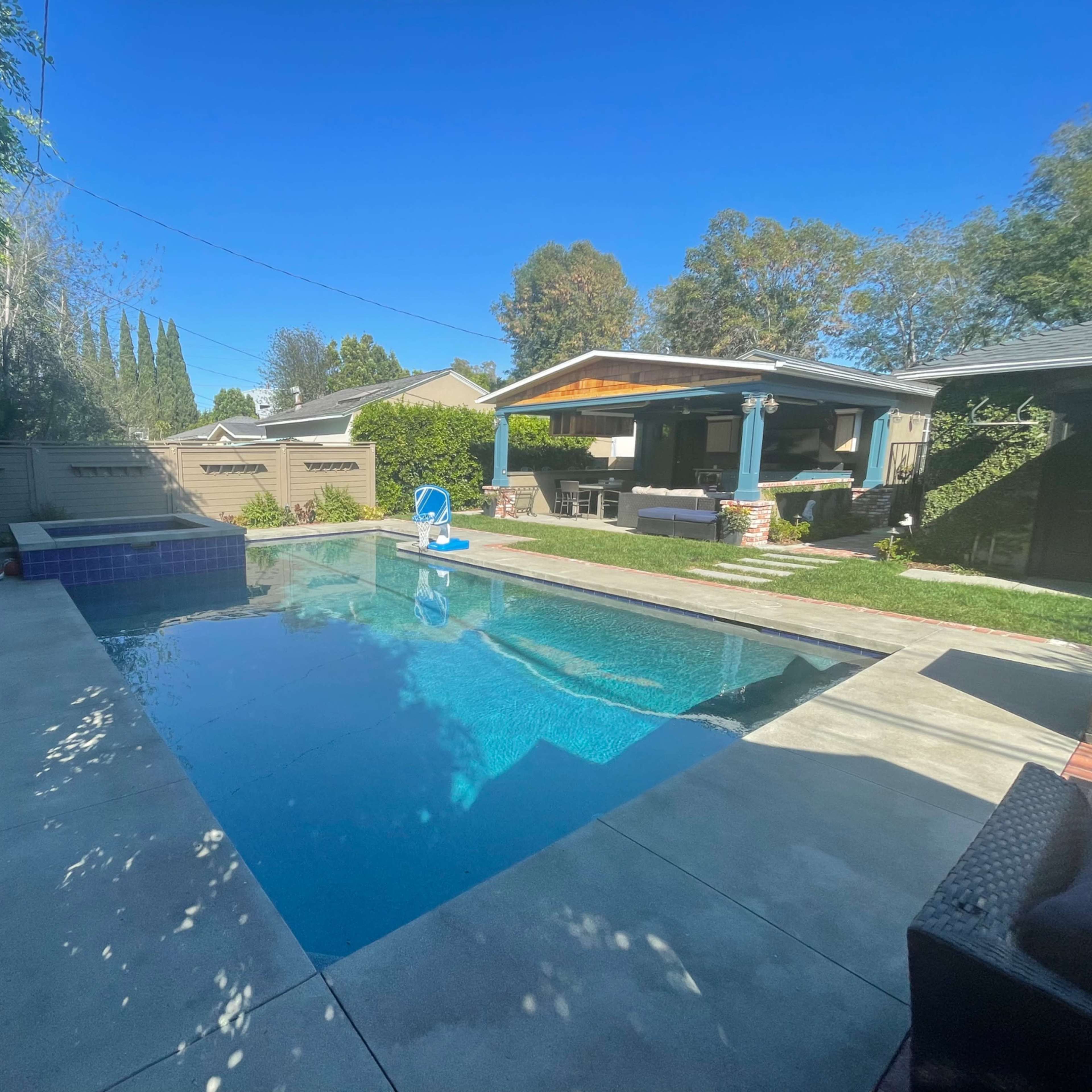 A clear swimming pool surrounded by a concrete patio and grass, with a shaded seating area and a spa on one side.