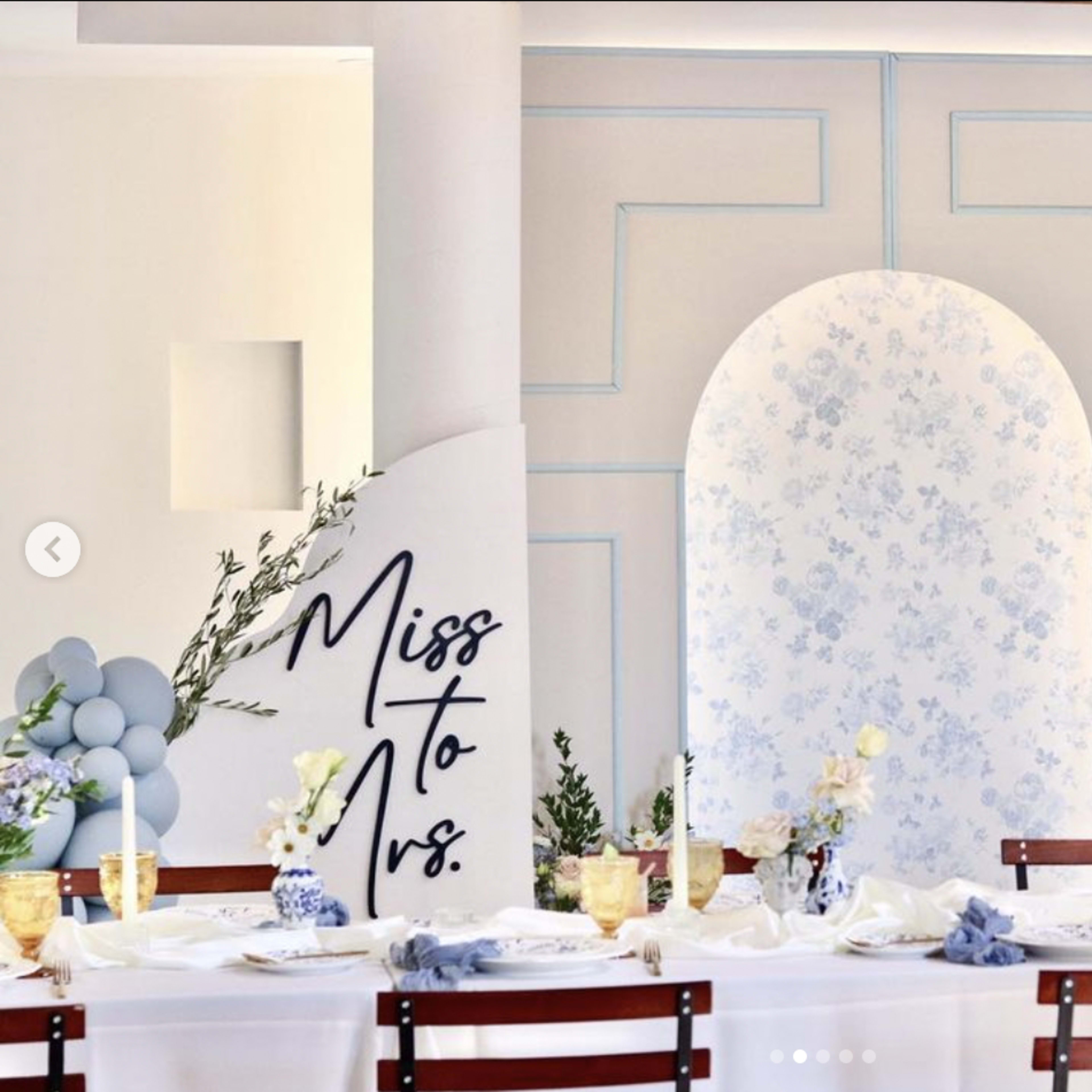 A decorated dining table is set against a white wall featuring floral patterns and an arch, with a sign that reads "Miss to Mrs." on the side.