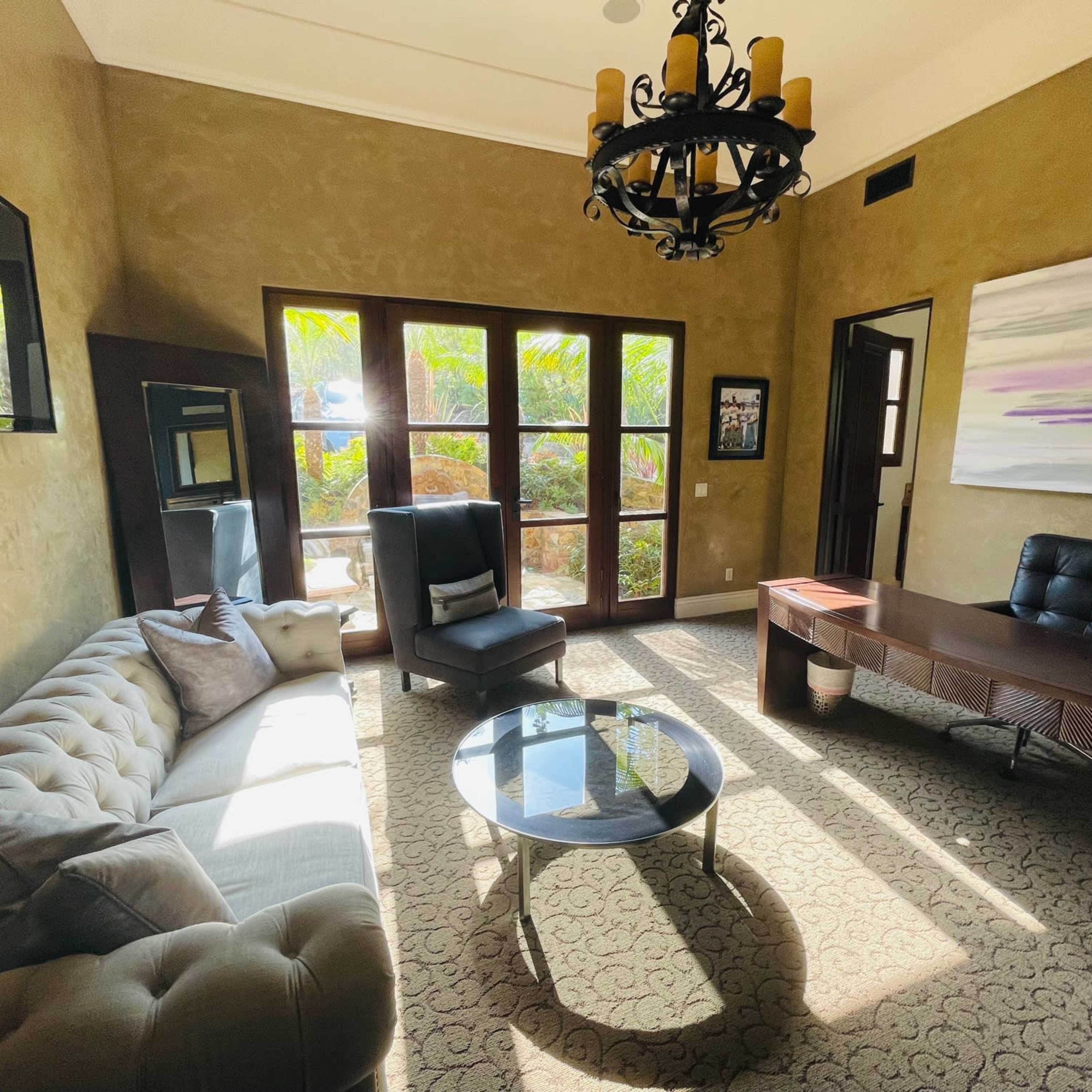 A well-lit office with a tufted sofa, a glass coffee table, a dark wooden desk, and large windows overlooking greenery.
