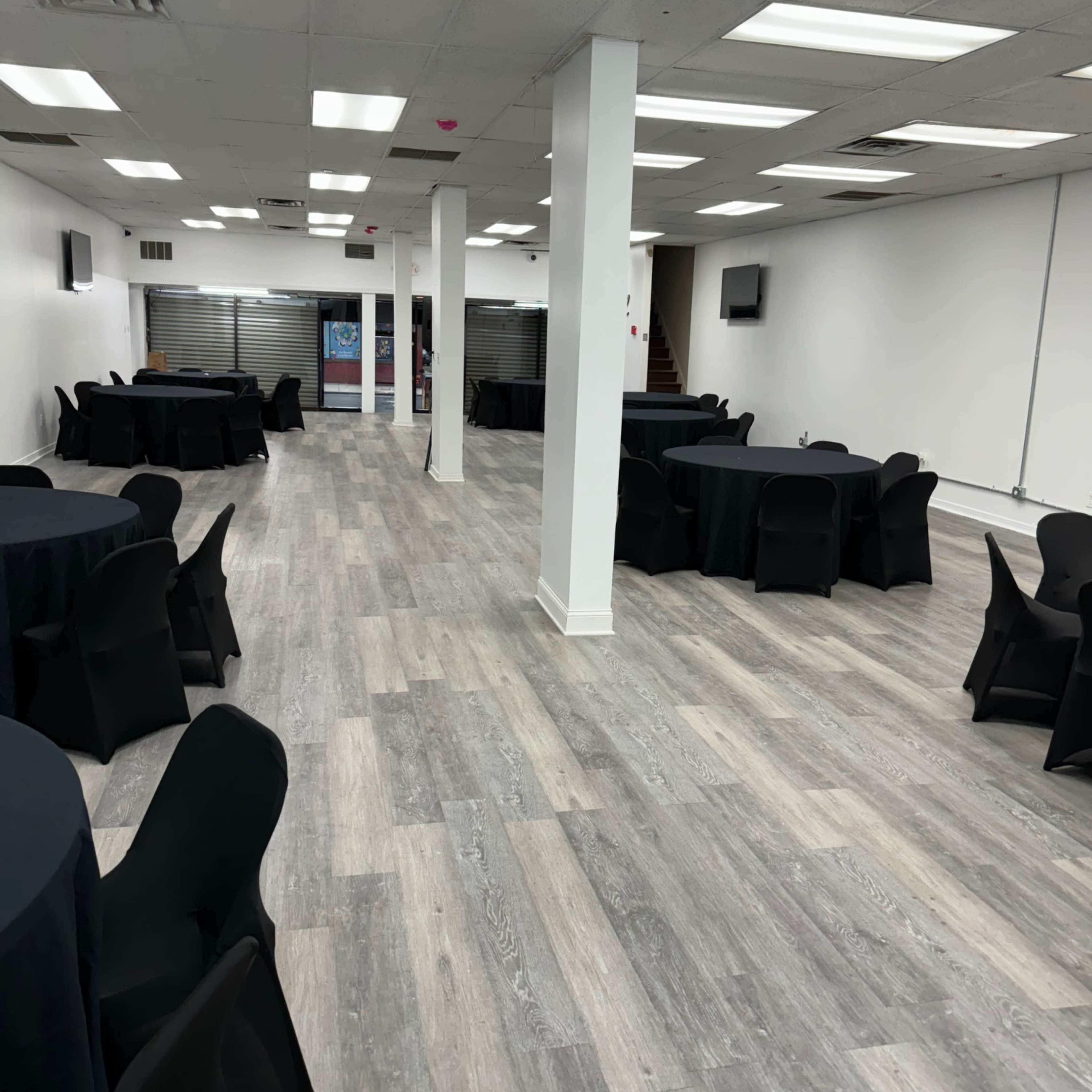 A spacious event room with black tablecloths covering round tables arranged throughout, and a light-colored wooden floor.