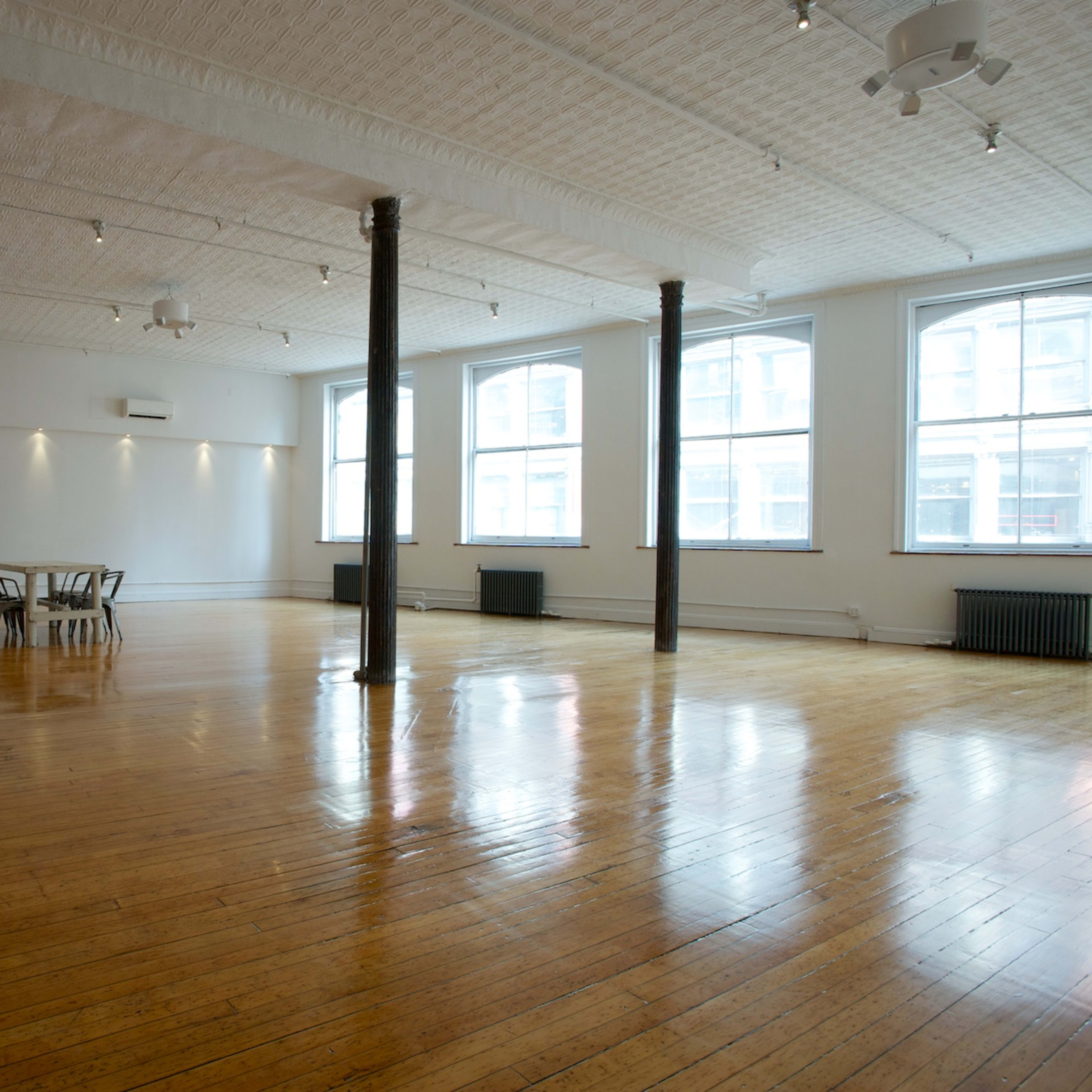 A spacious, empty room with wooden flooring, large windows, and exposed structural columns.