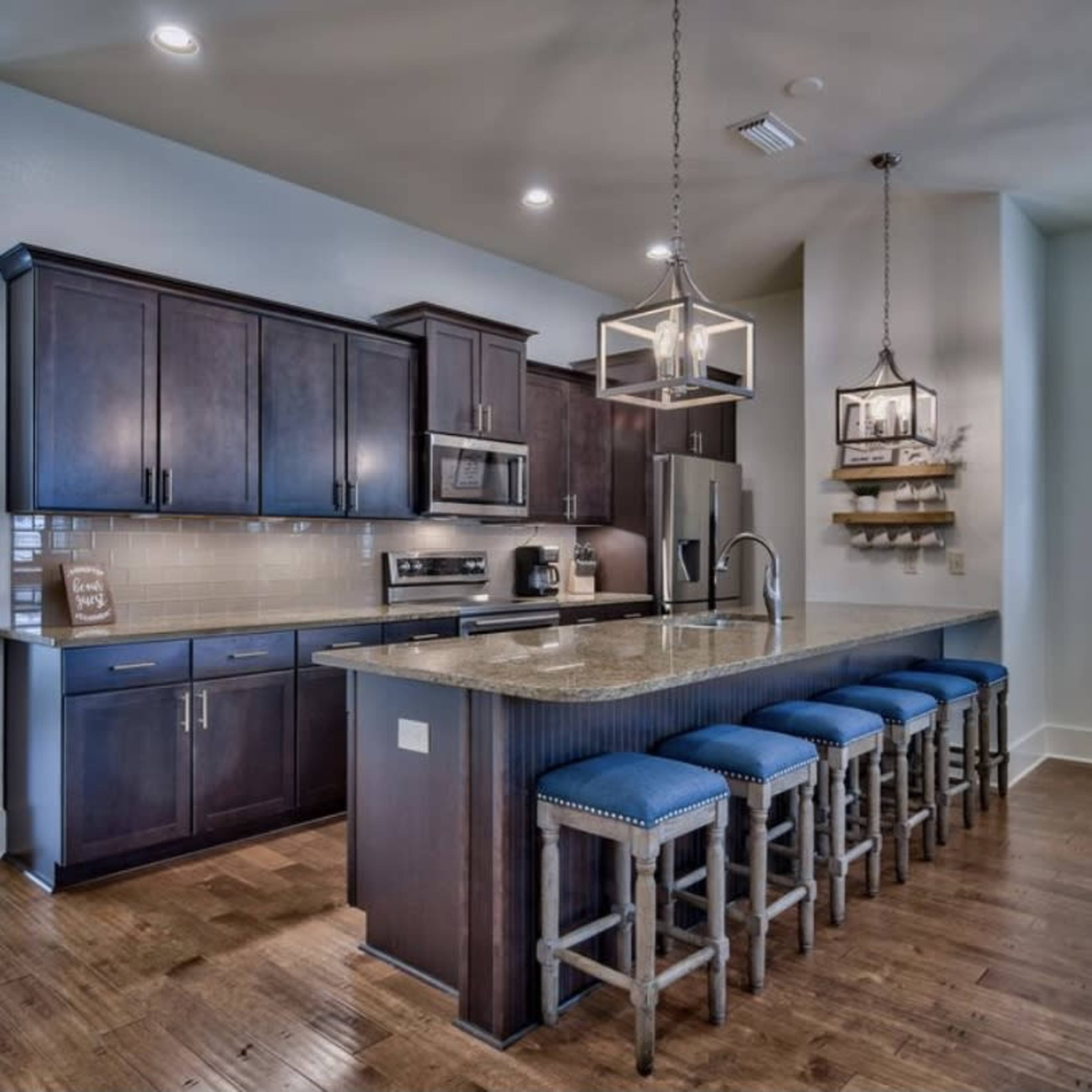 A modern kitchen with dark wooden cabinets, a large granite countertop, and blue upholstered counter stools arranged in front of it.