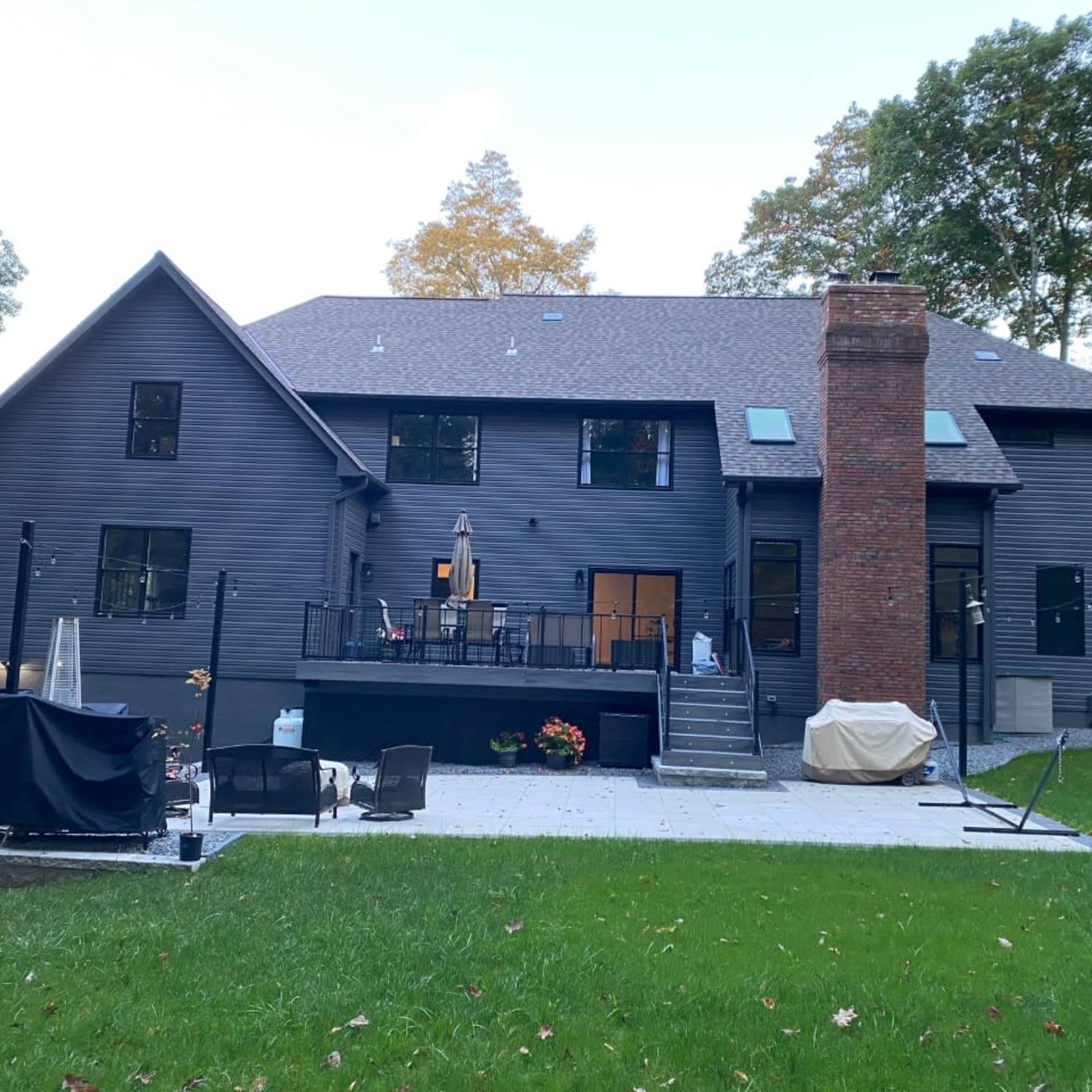 A modern two-story house with a dark exterior features a large patio, outdoor seating area, and a chimney.