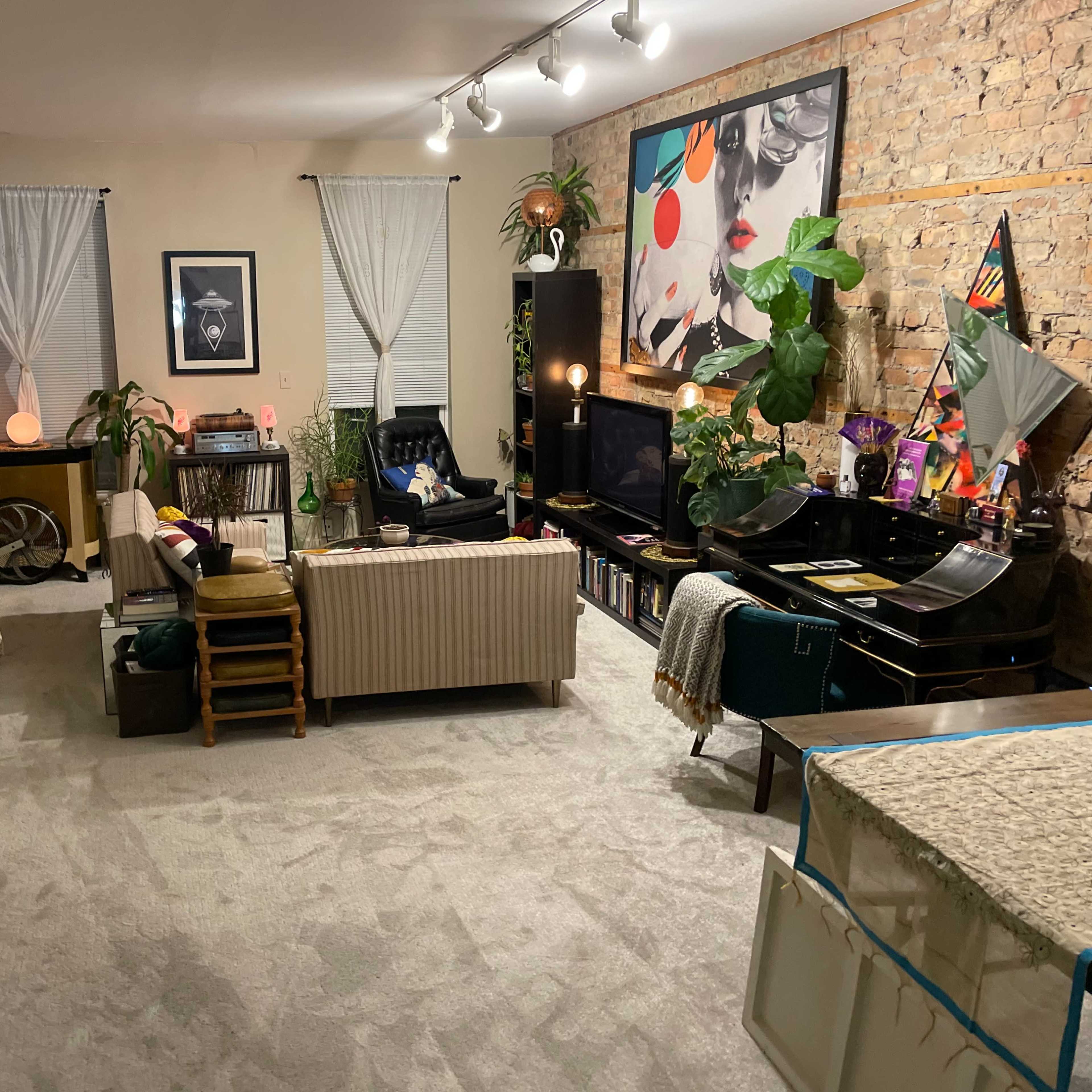 A cozy living room features a mix of textiles, plants, and furniture arranged around a central seating area, with exposed brick walls and soft lighting.