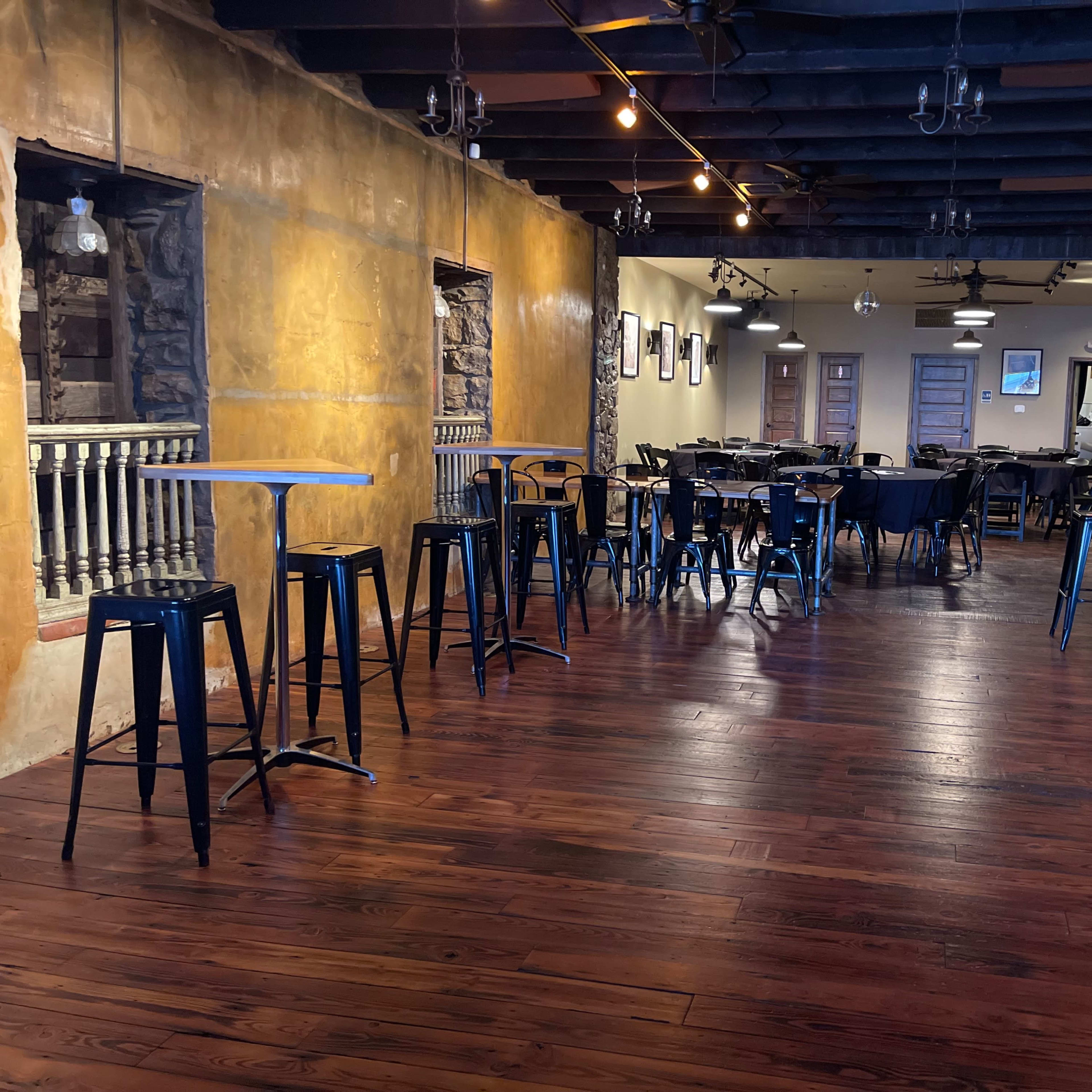 A spacious restaurant interior featuring dark wooden floors, a bar with high stools, and tables set up for dining.
