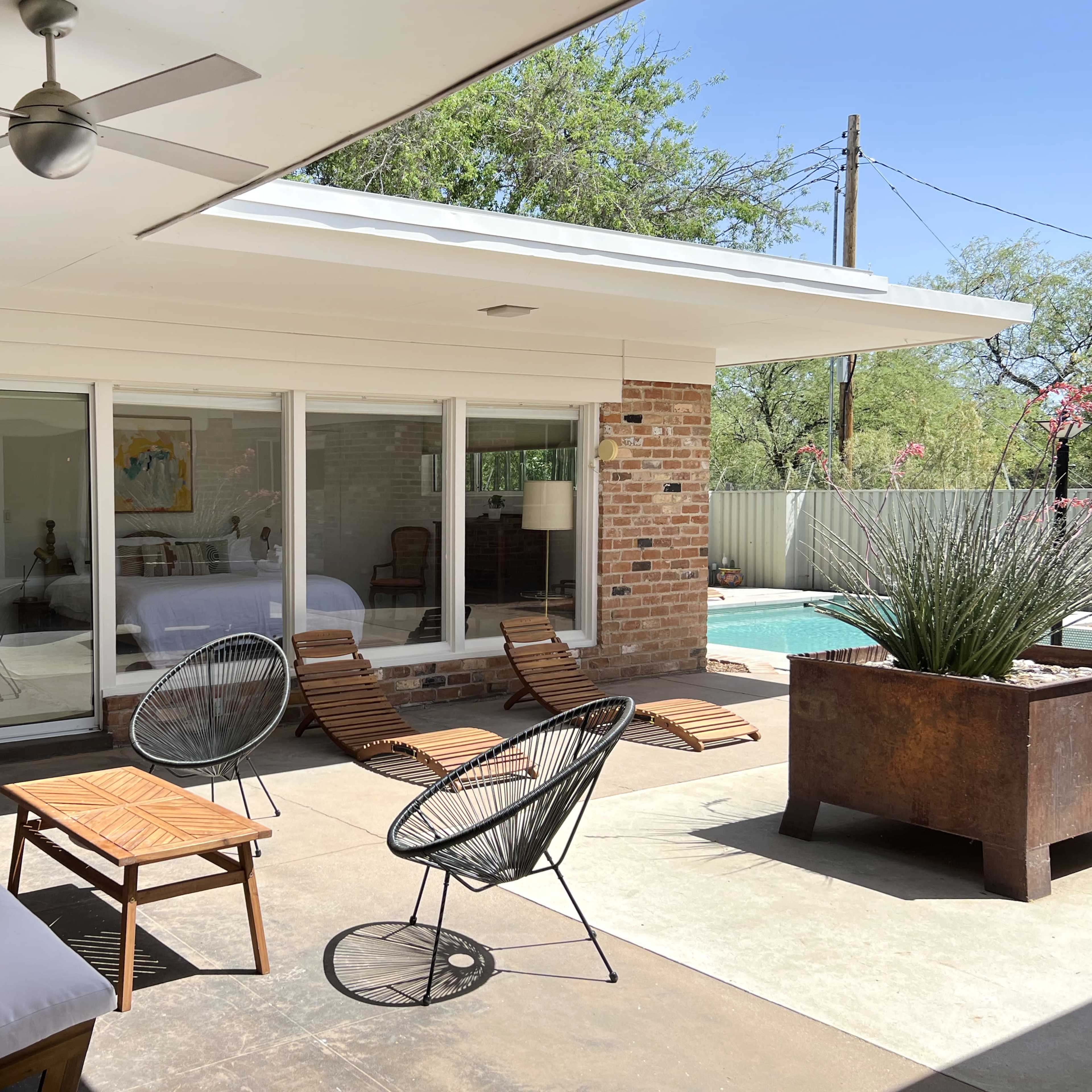 A patio area with a seating arrangement consisting of a white sofa, wooden coffee table, and black chairs, overlooking a pool and a bedroom area through large glass doors.