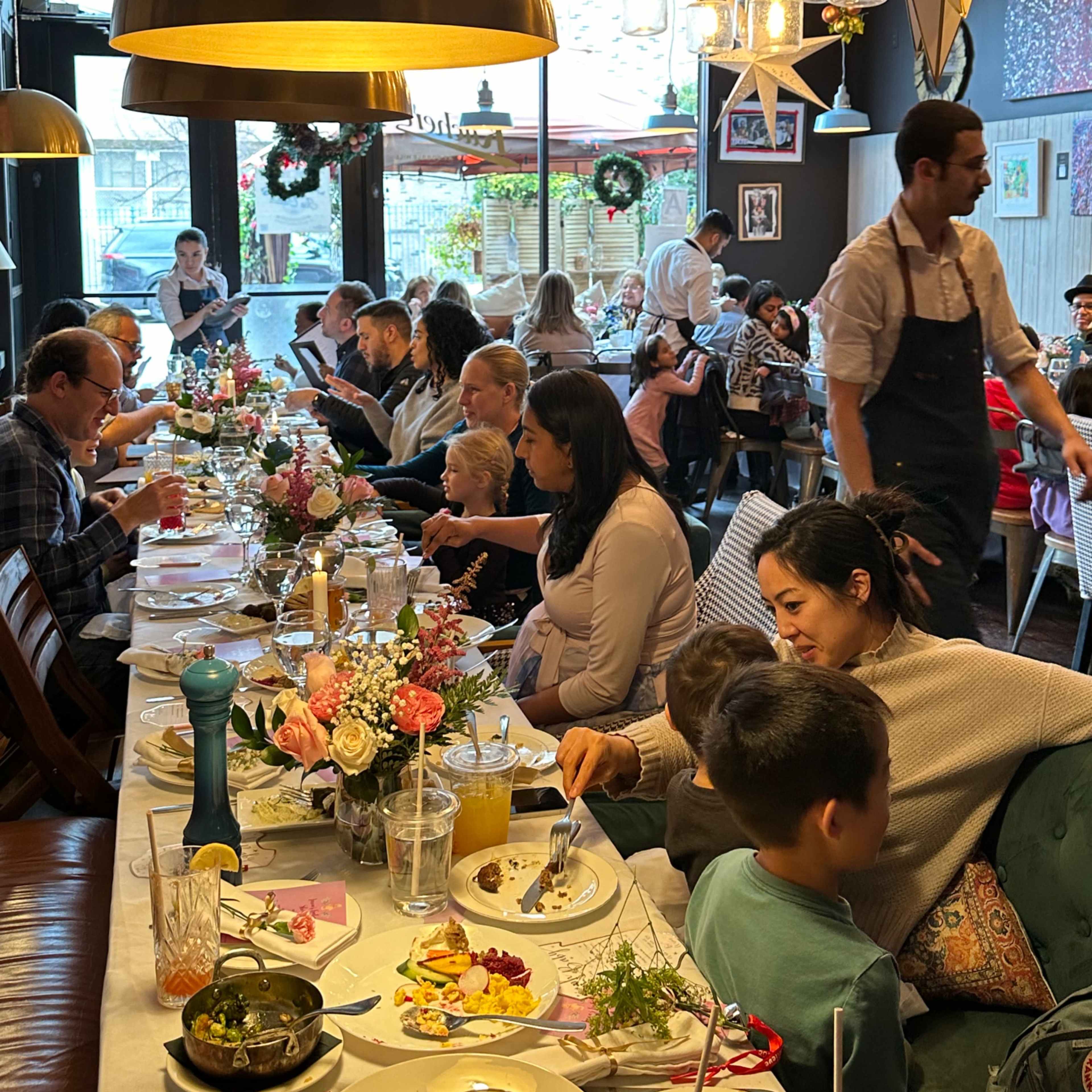 A bustling restaurant scene shows diners enjoying meals at a long table adorned with flowers, while a server attends to guests in a lively atmosphere.