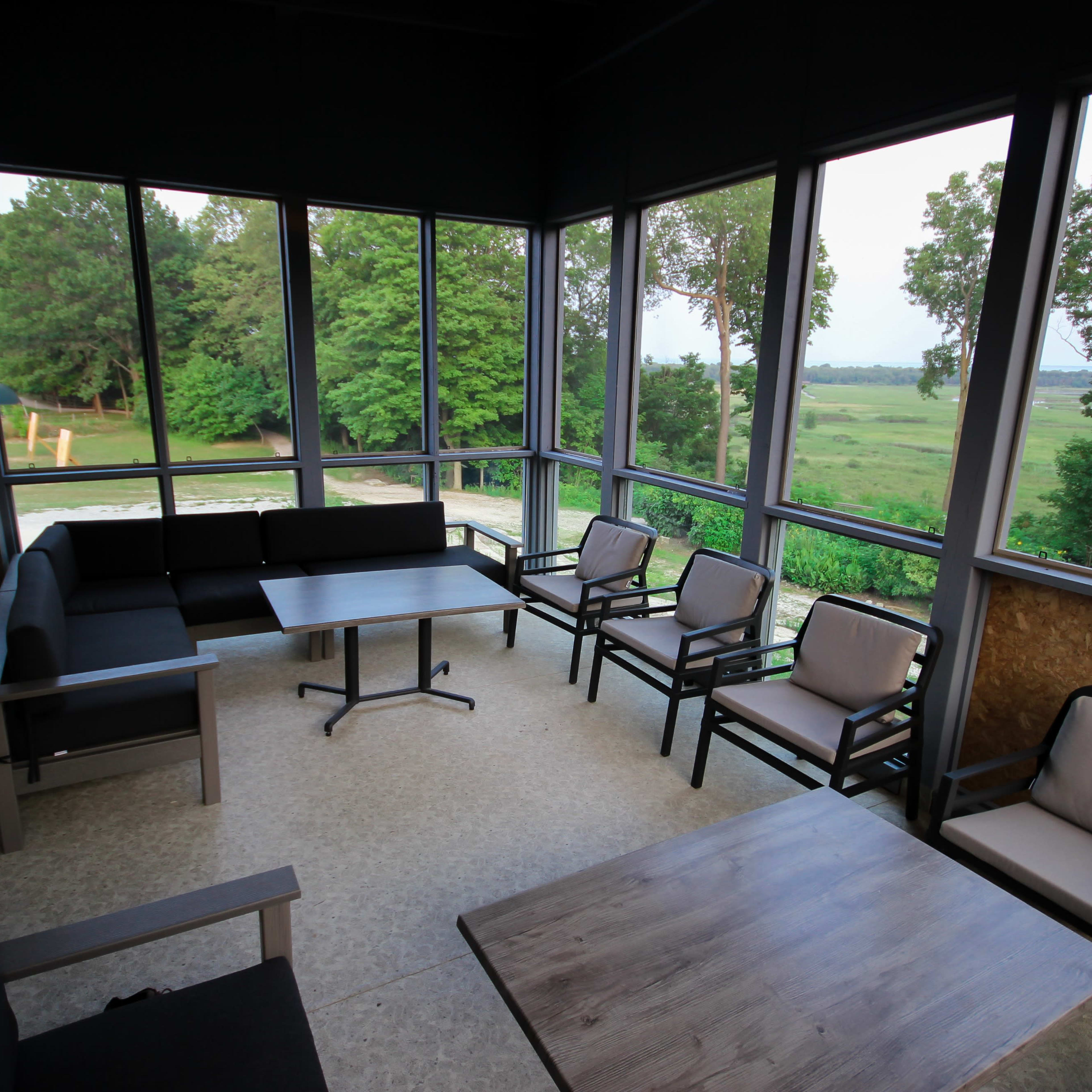 A screened-in porch with a seating area comprising a table and multiple chairs, overlooking a green landscape through large windows.