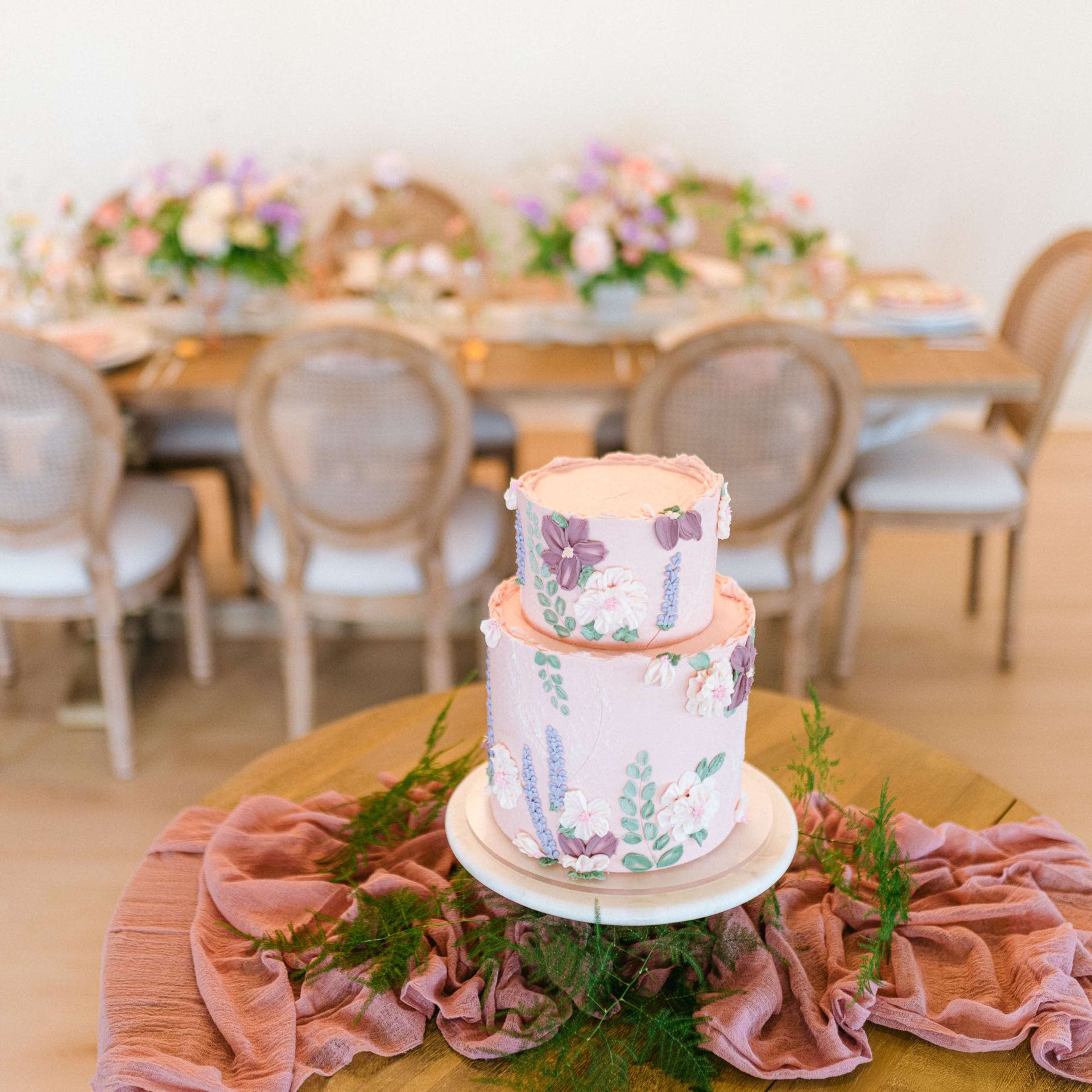 A two-tiered pink cake adorned with floral decorations is set on a table in front of a dining area with a wooden table and chairs surrounded by floral centerpieces.