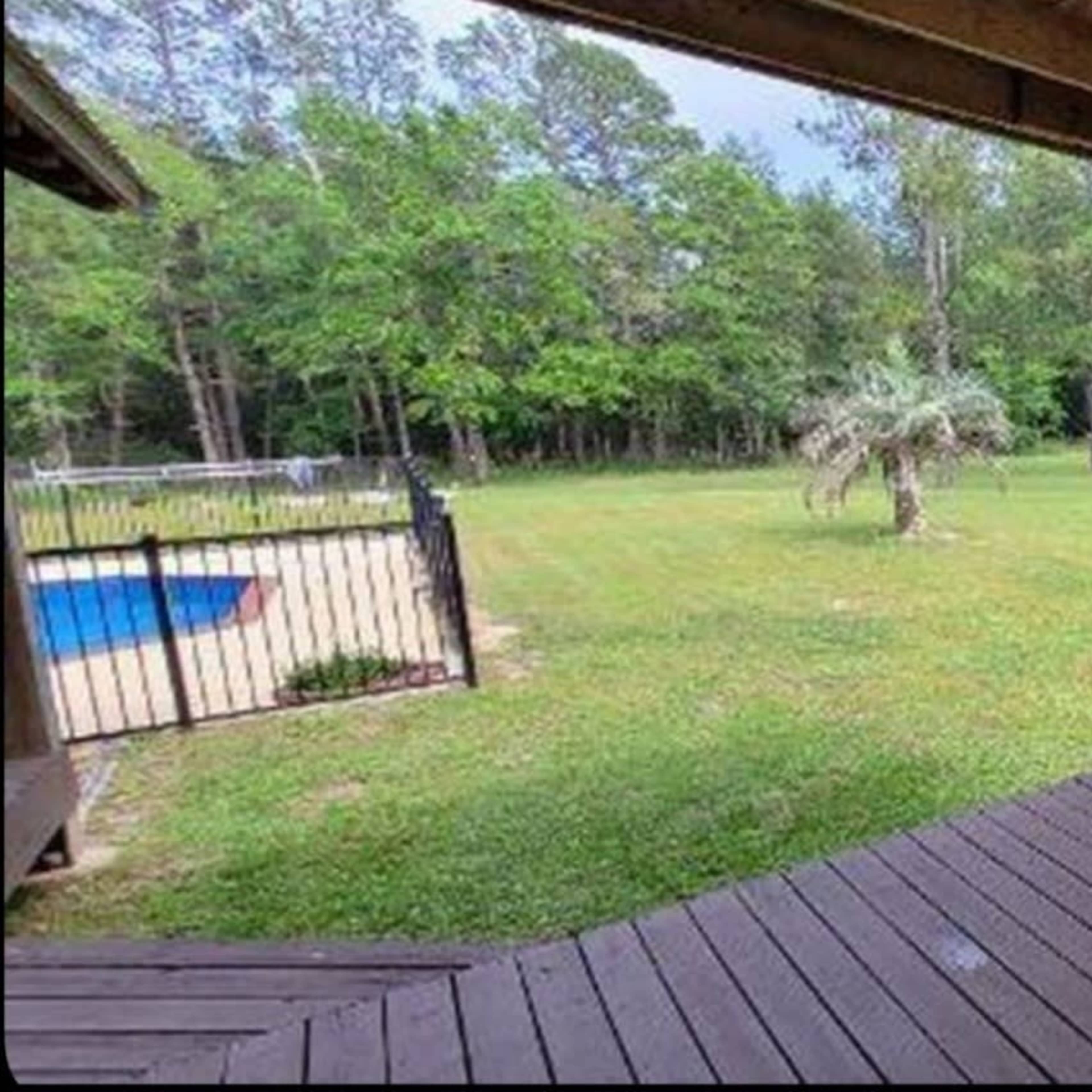 A hammock is suspended from a porch overlooking a grassy yard with trees and a pool in the background.