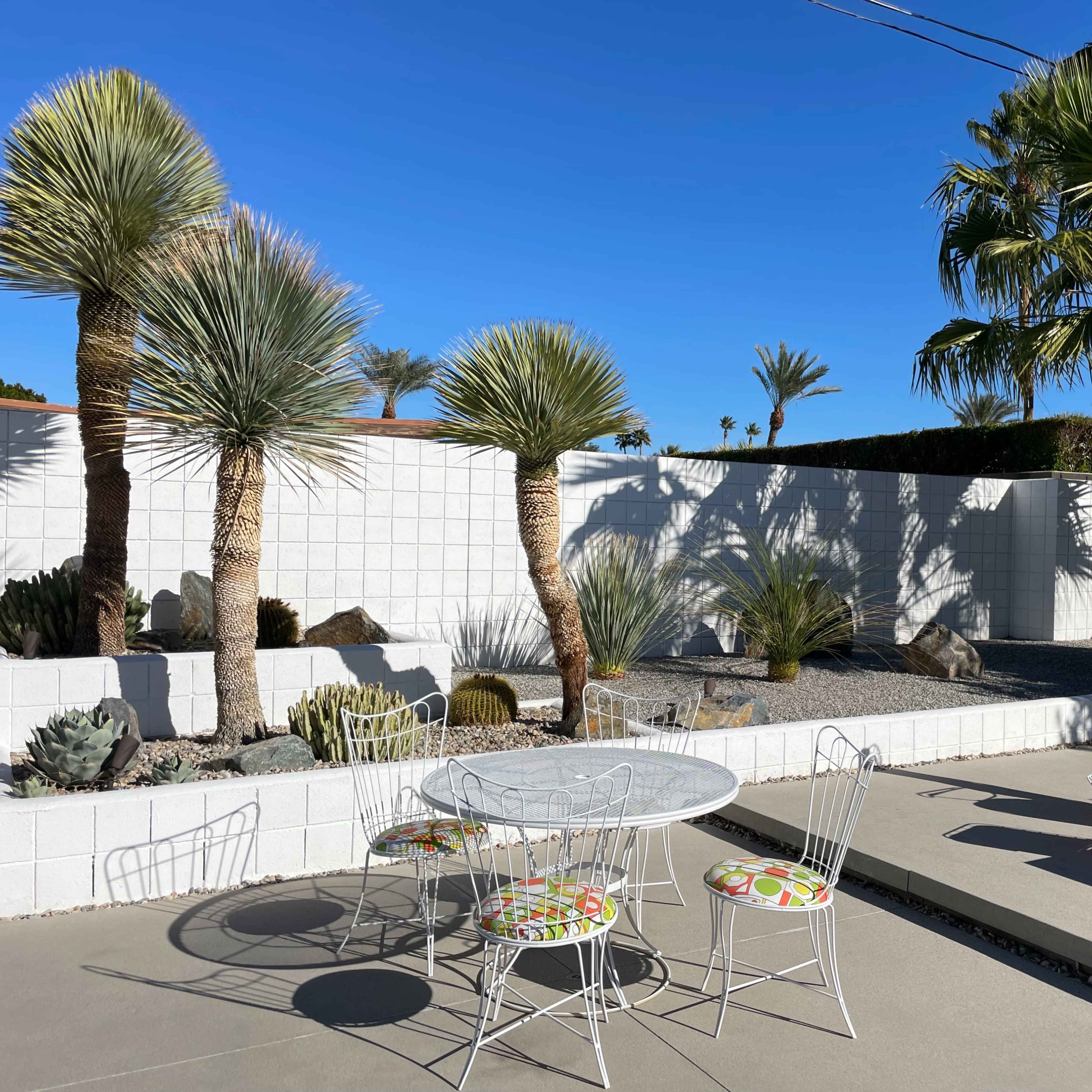 A patio area features a white wire table surrounded by four chairs, with a colorful lounge chair nearby, set against a backdrop of palm trees and a low white wall.