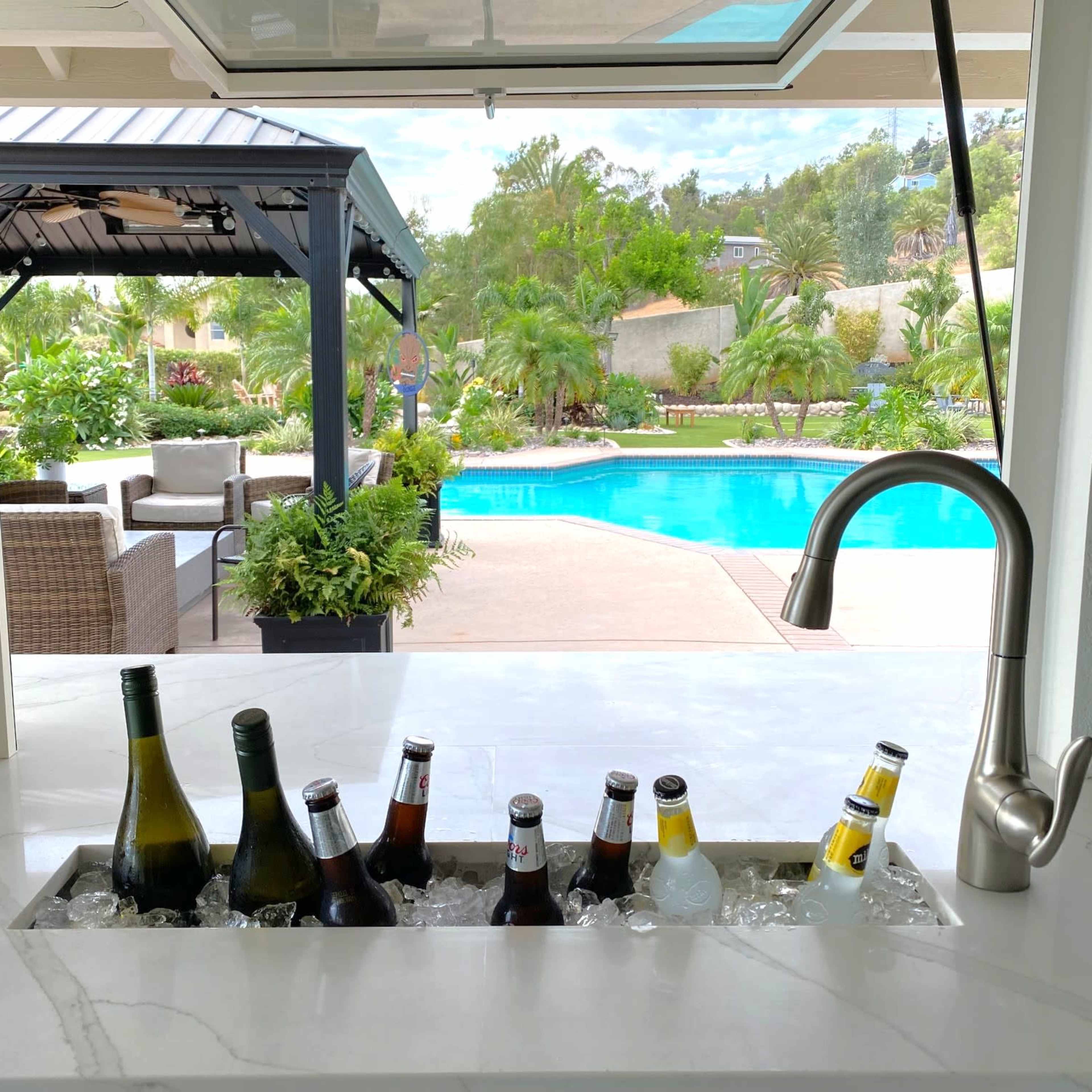 An outdoor kitchen area features a sunken ice bucket filled with various bottles, overlooking a swimming pool surrounded by lush landscaping.