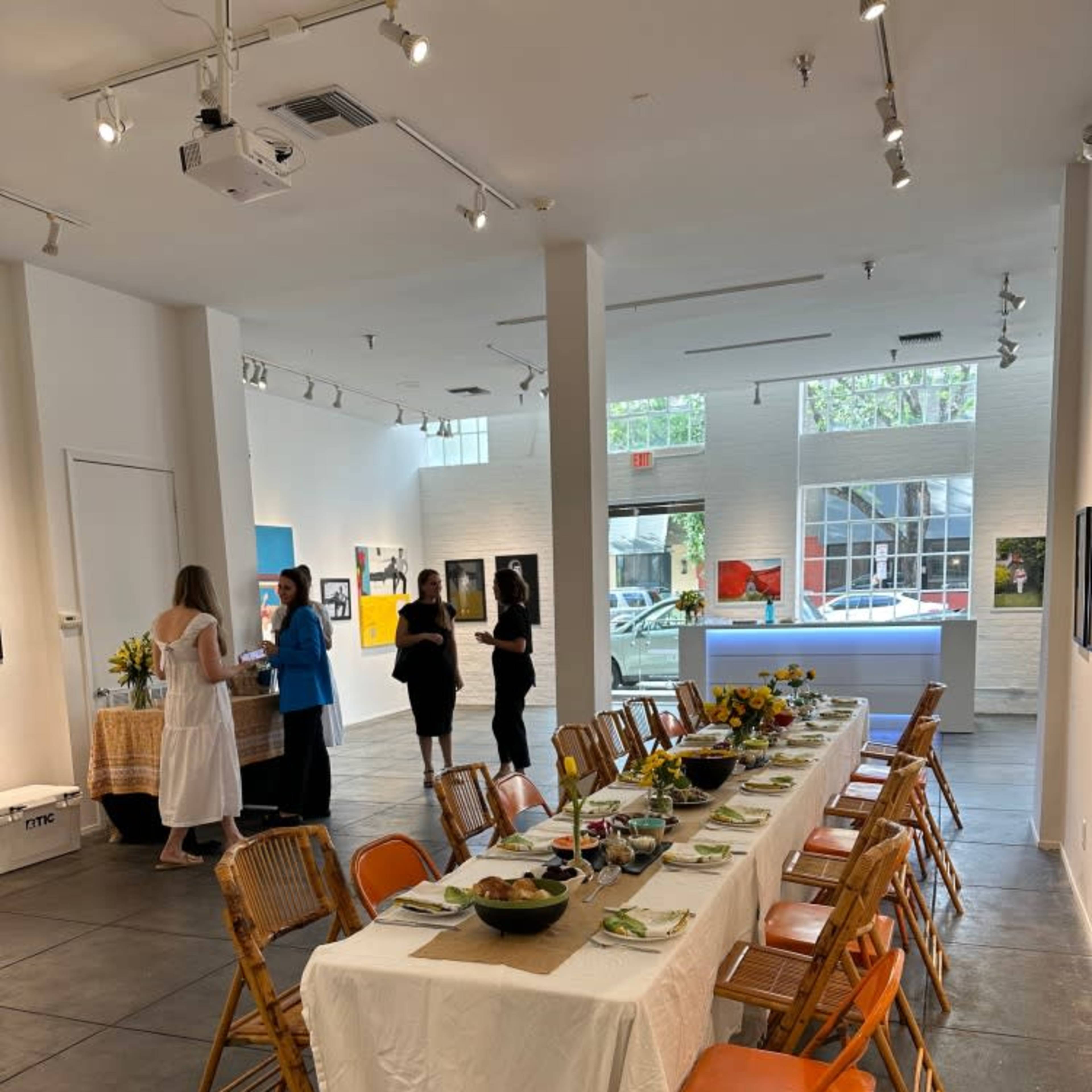 A long dining table is set in an art gallery, with guests mingling and a display of artworks on the walls.