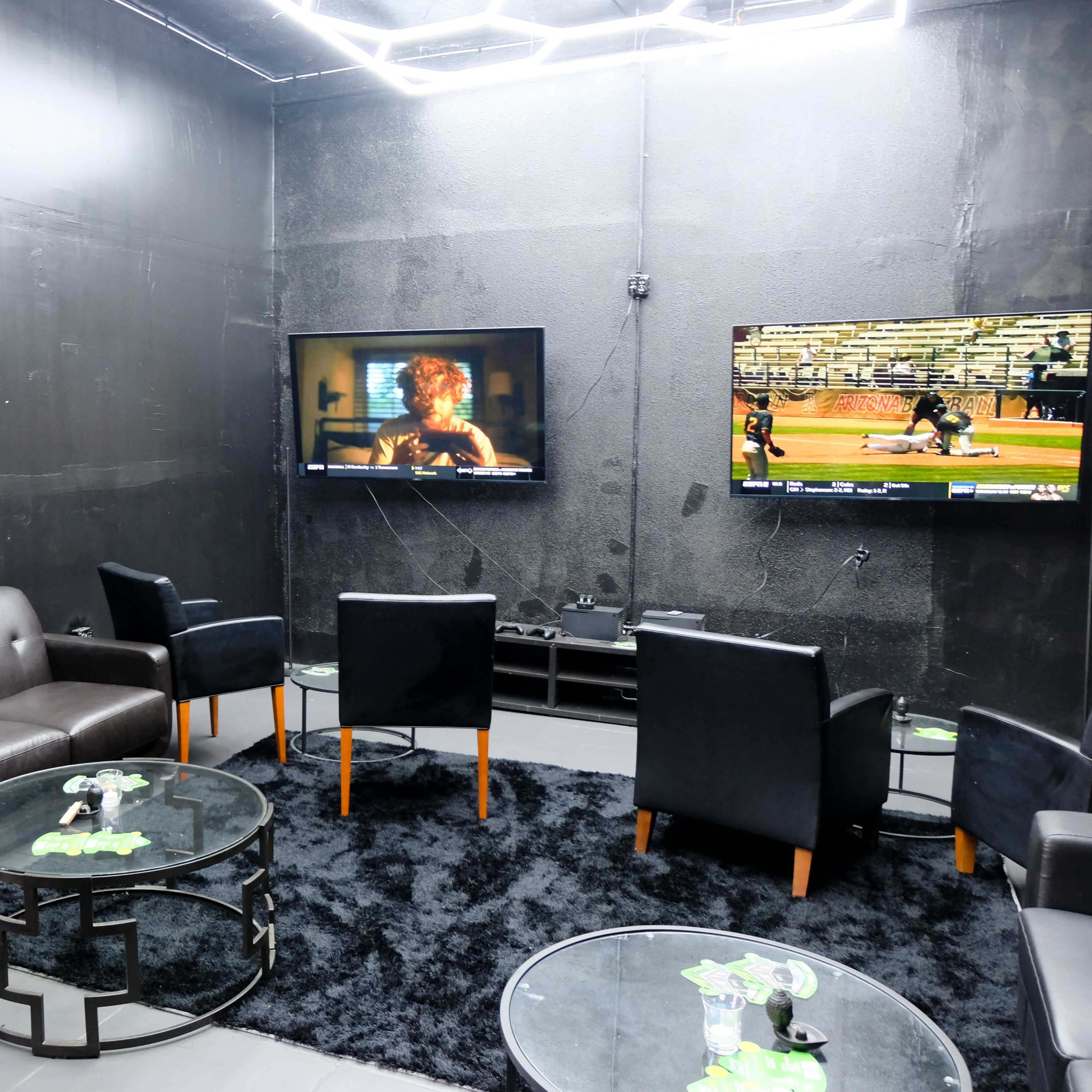 A dimly lit lounge area with black walls, a black sofa, several black chairs, and two televisions displaying different programs.