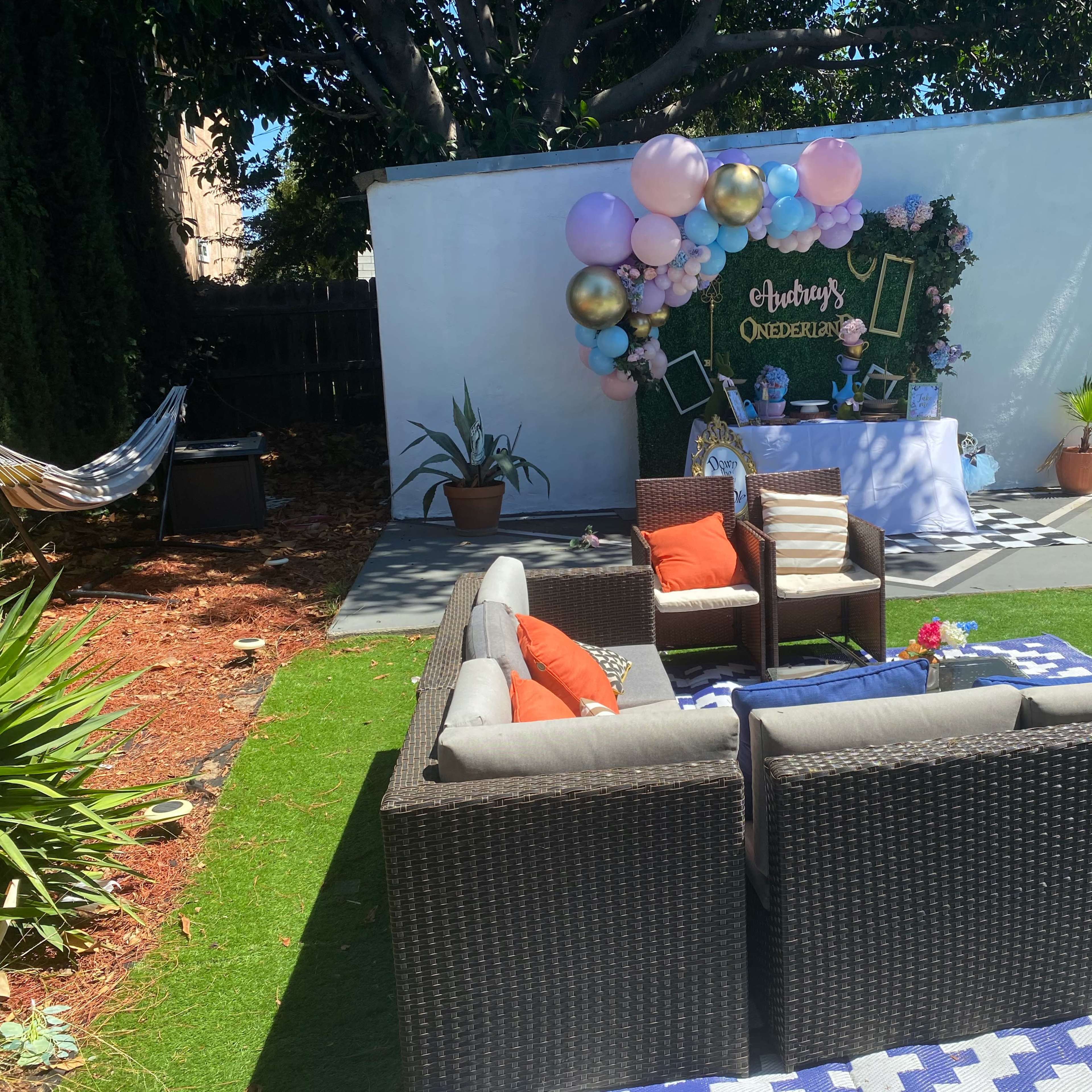 An outdoor party setup with a seating area consisting of a brown wicker couch, colorful pillows, a decorated wall with balloons, and a bouncy castle in the background.