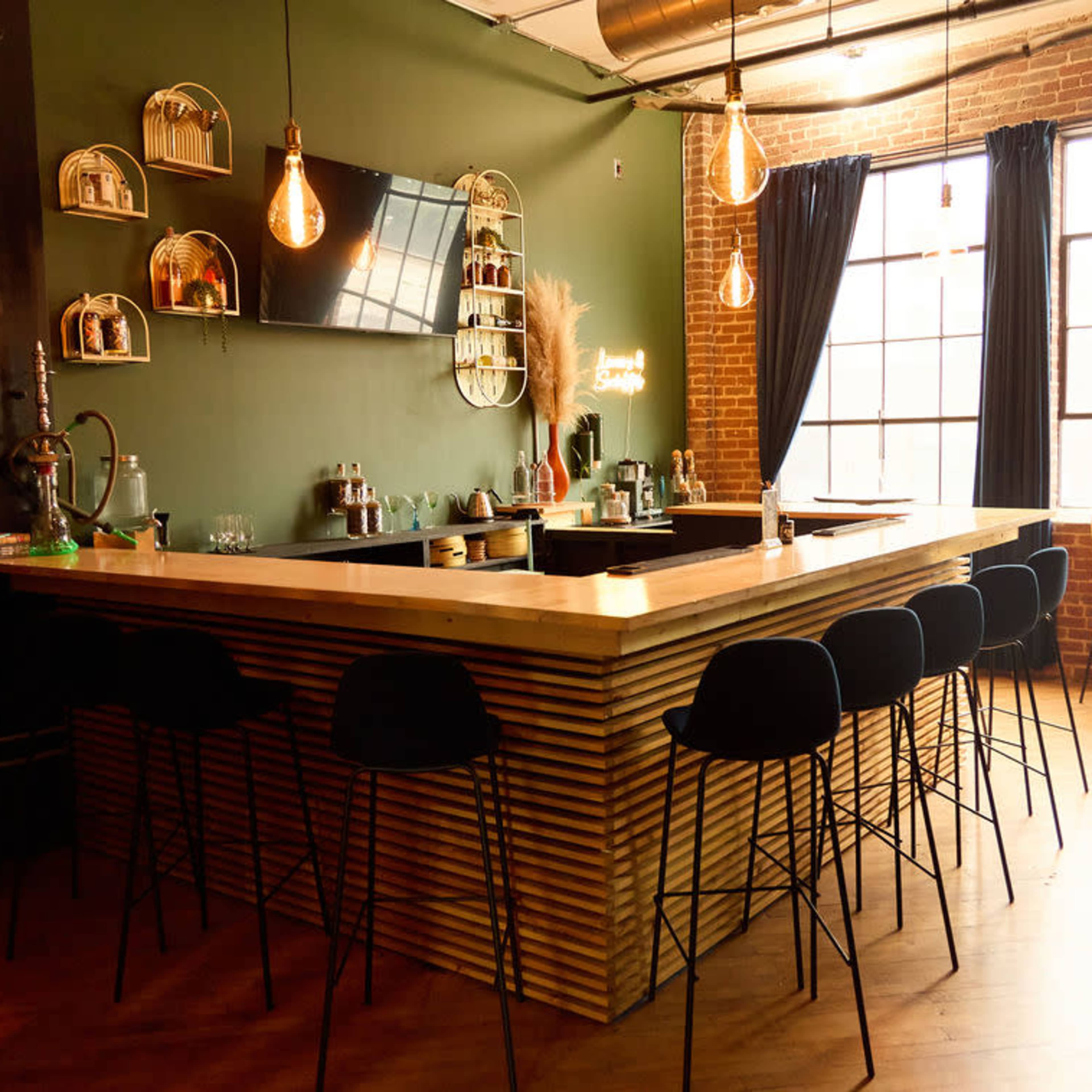 A modern bar area features a wooden counter, black stools, and large windows framed by dark curtains in a space with exposed brick walls.