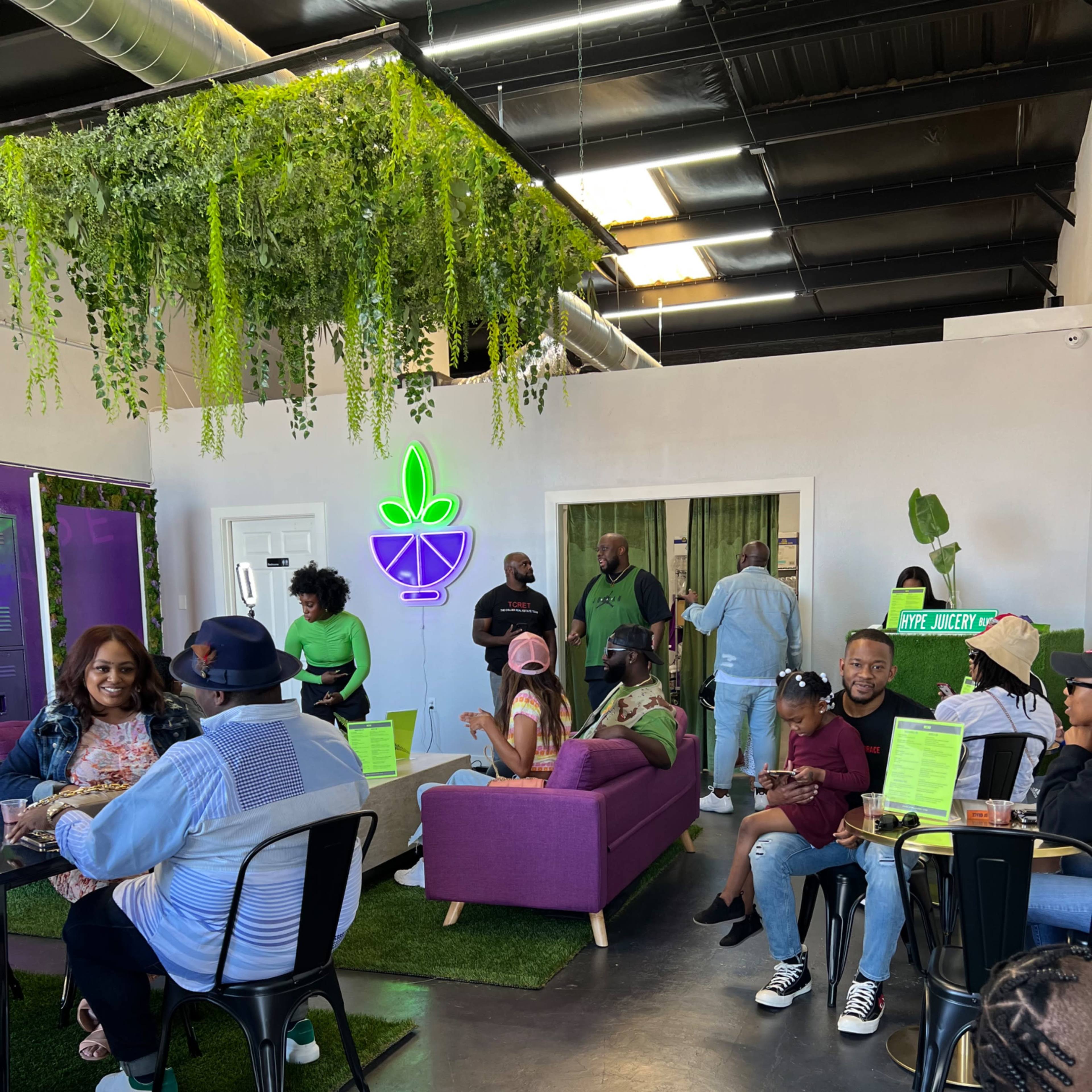 A vibrant indoor space features a green-themed decor with plants, seating areas, and people engaging in conversation and dining.