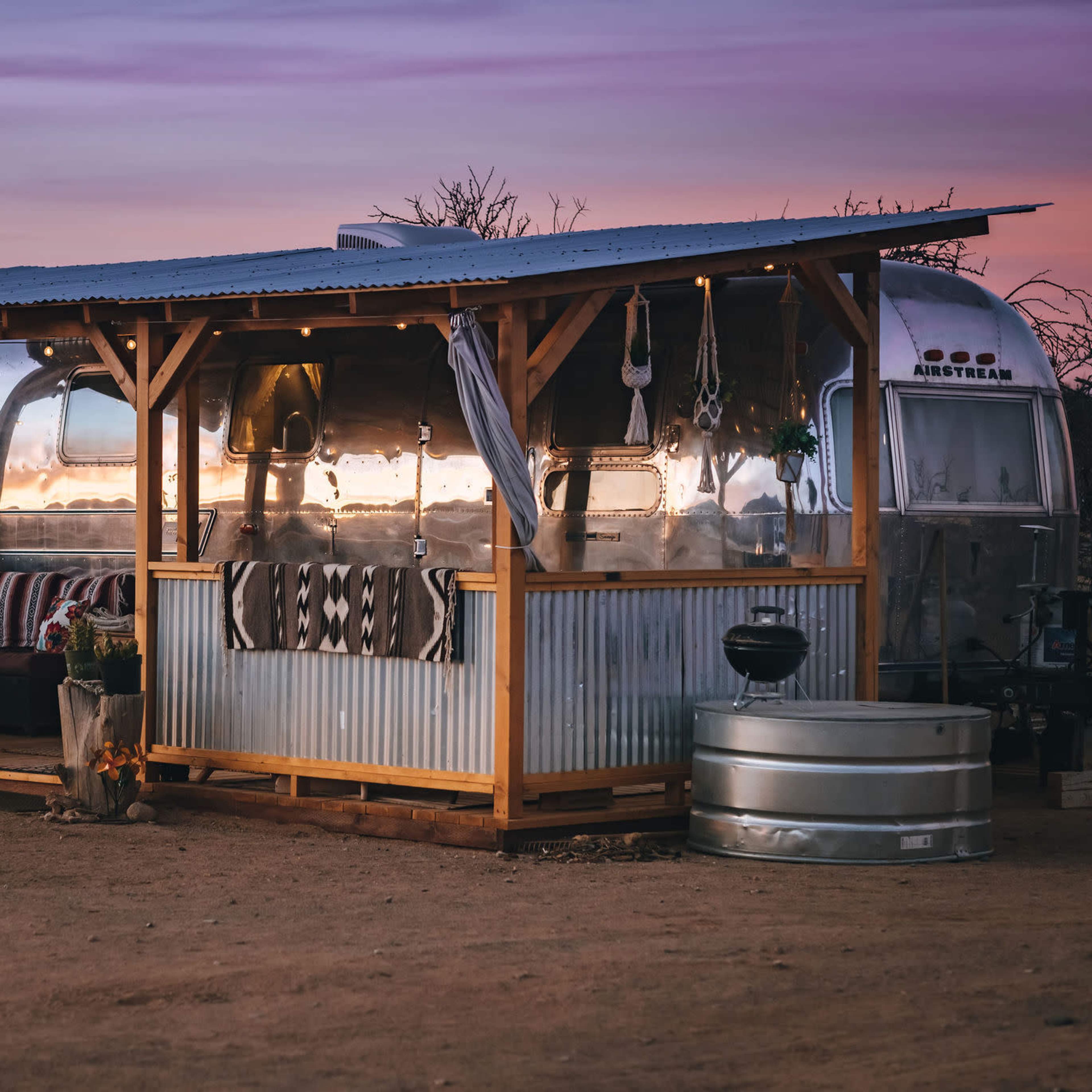 A silver Airstream trailer is set on a desert landscape, surrounded by a wooden porch with seating and decorative plants, as the sky displays hues of purple and orange at sunset.