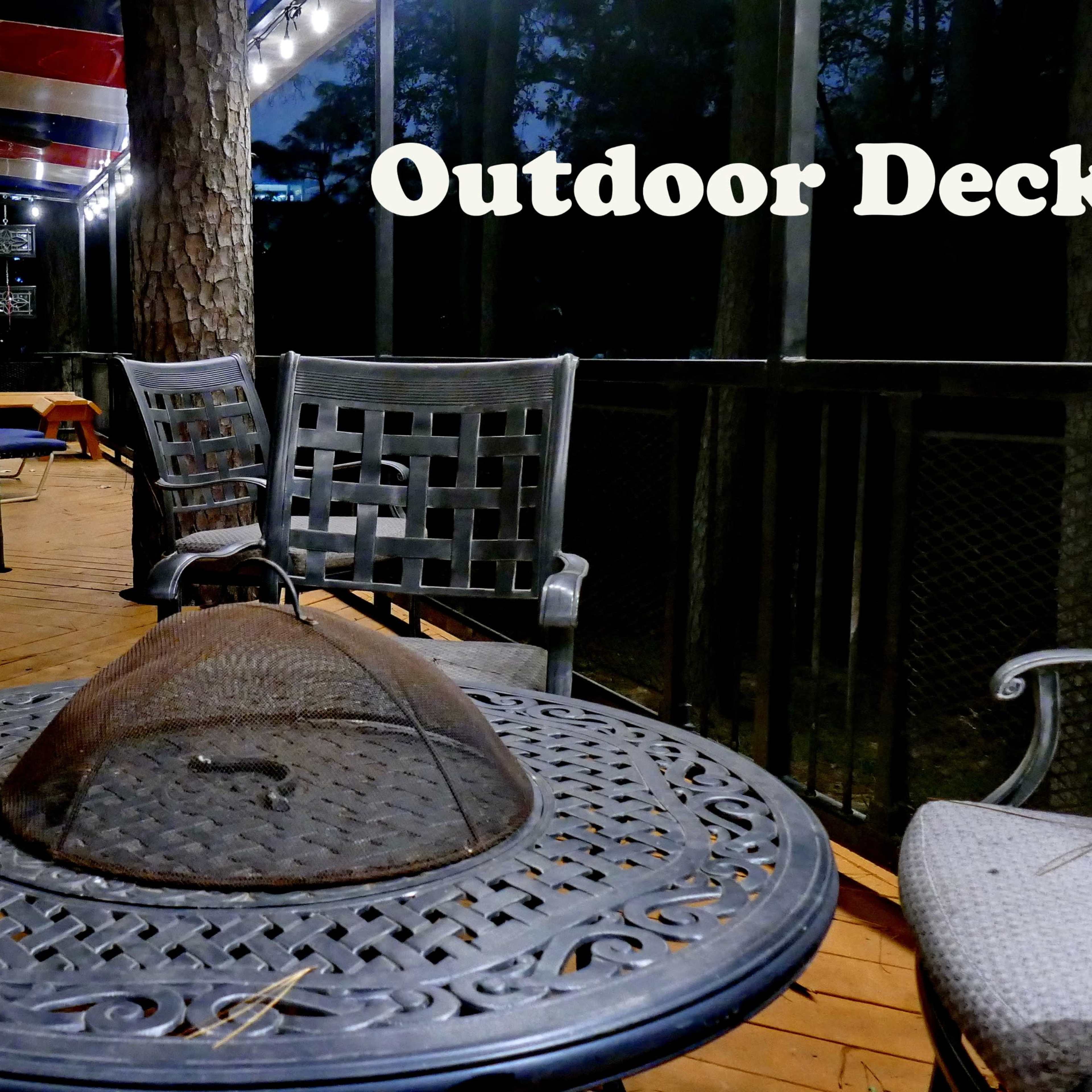 An outdoor deck features a round table with a mesh fire pit cover surrounded by four metal chairs.