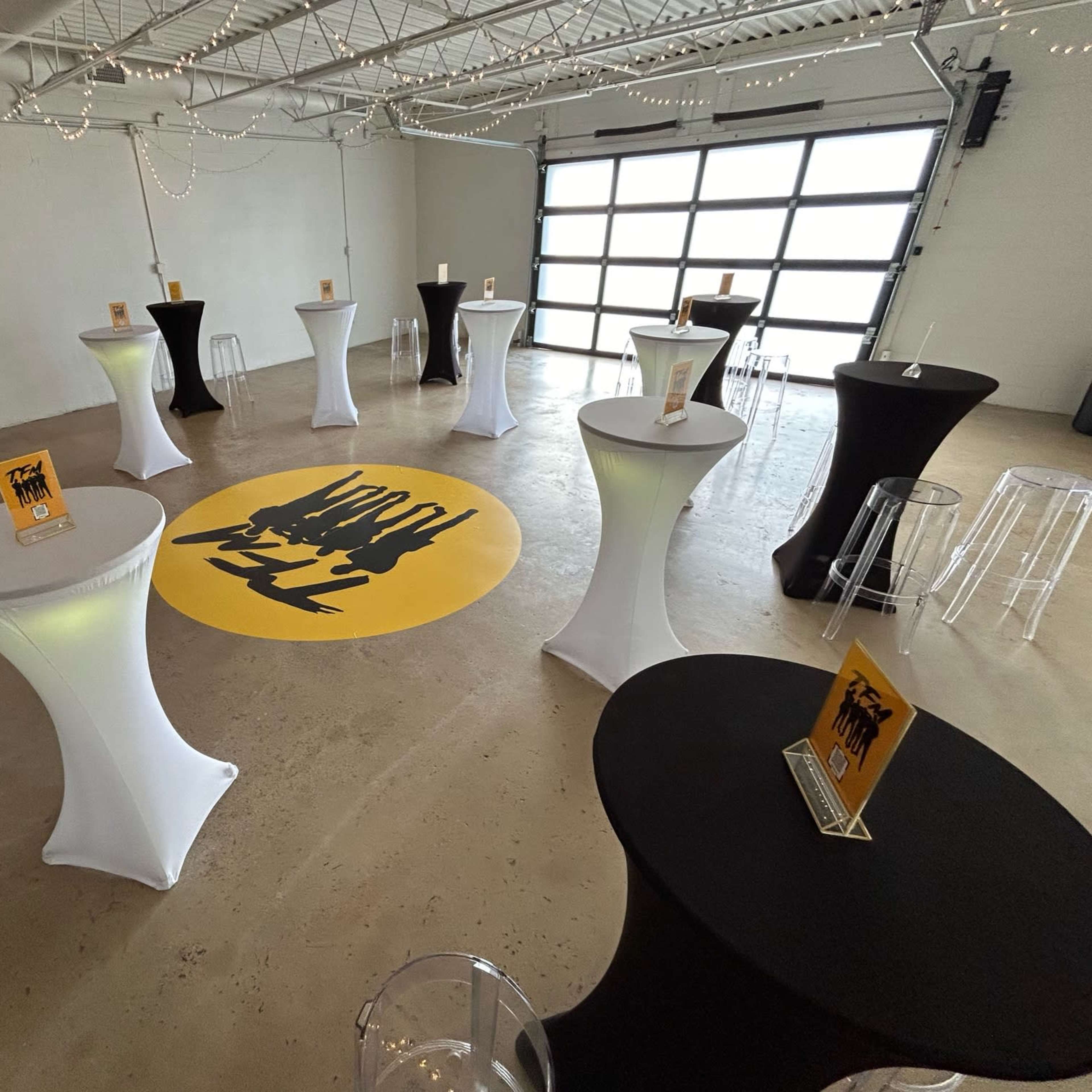A spacious indoor venue with a polished concrete floor and multiple tall tables adorned with table covers in white and black, along with decorative centerpieces.