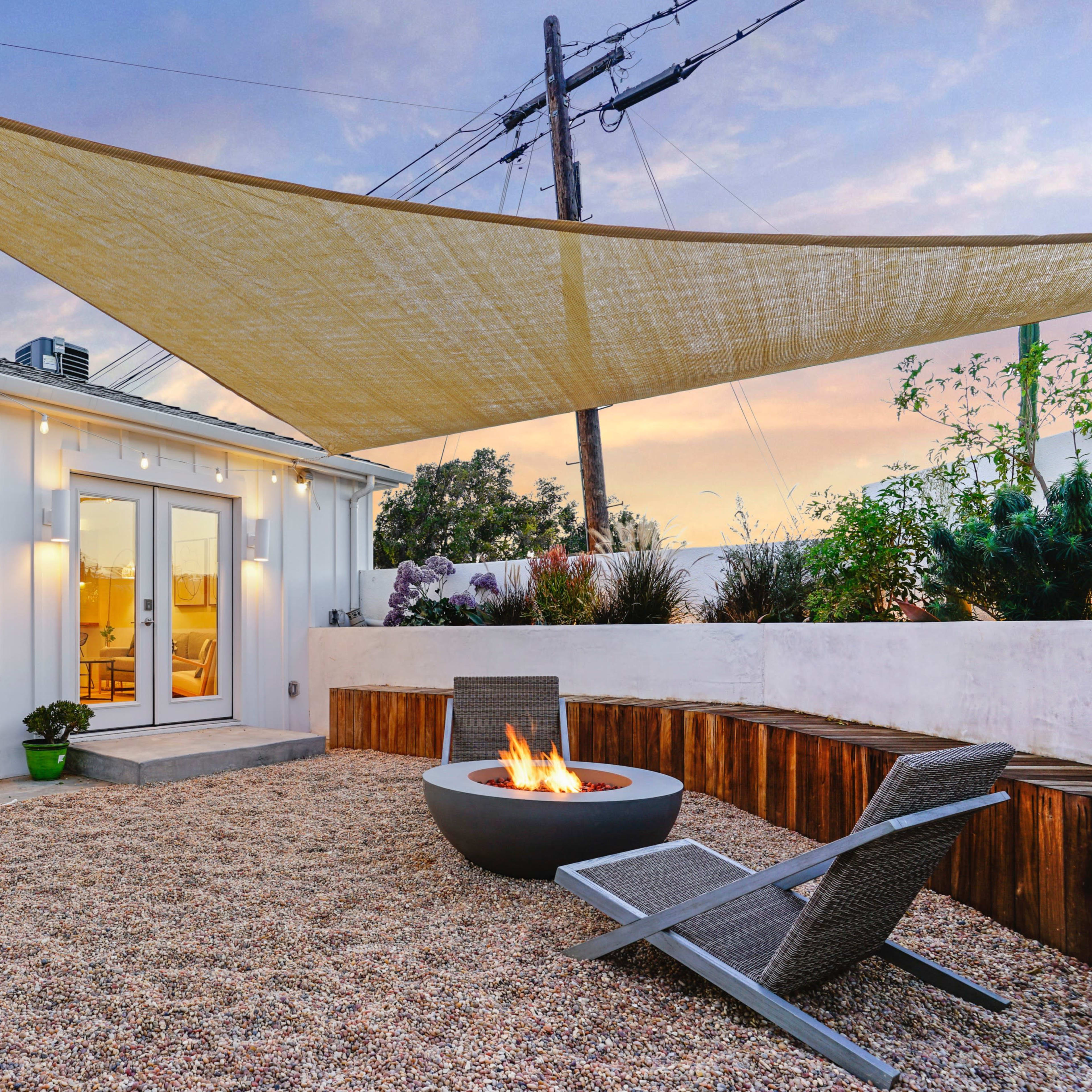 A modern outdoor space features a fire pit surrounded by gravel, lounge chairs, and a shaded area with a fabric canopy.