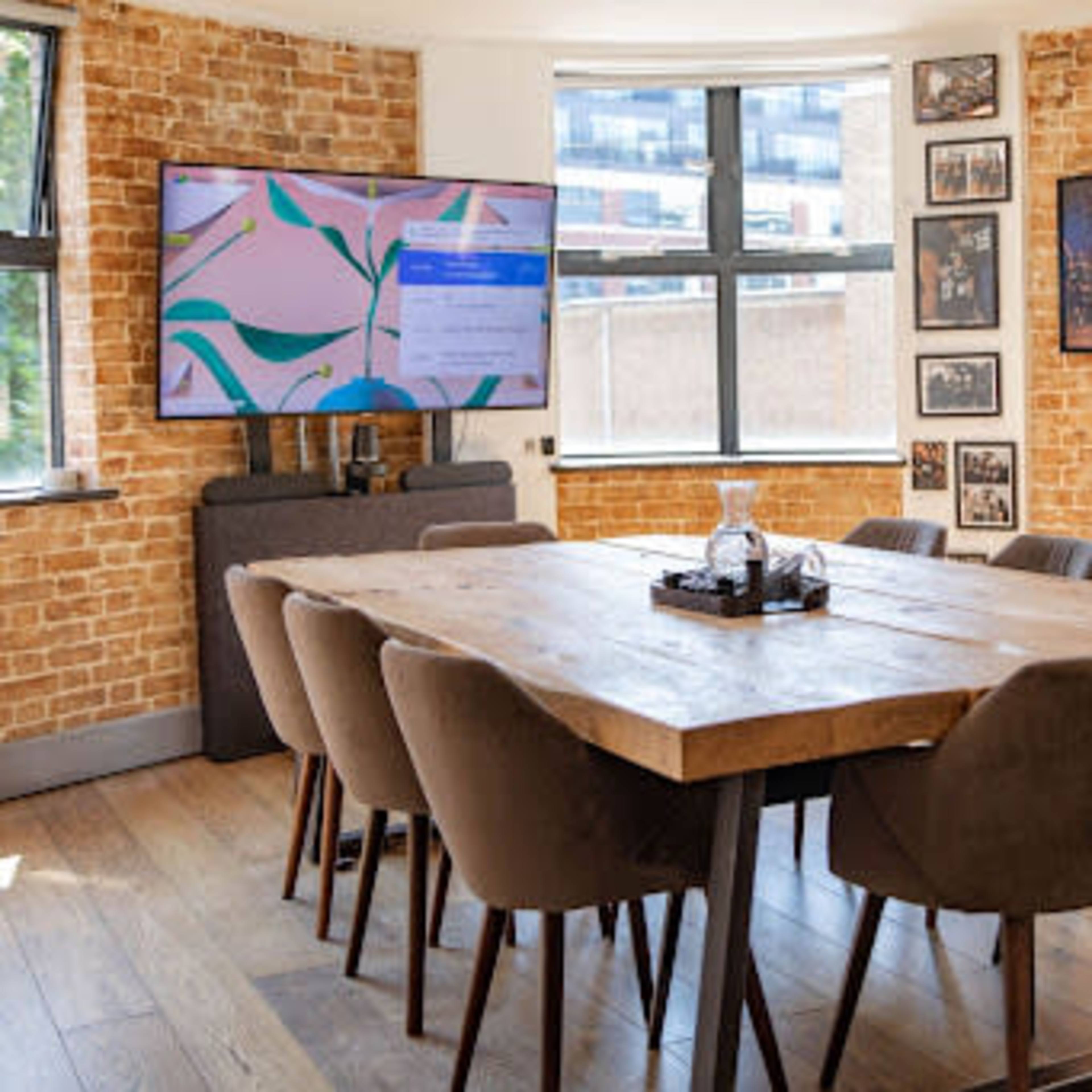A modern conference room features a large wooden table surrounded by upholstered chairs, with a television mounted on the wall and brick accents.