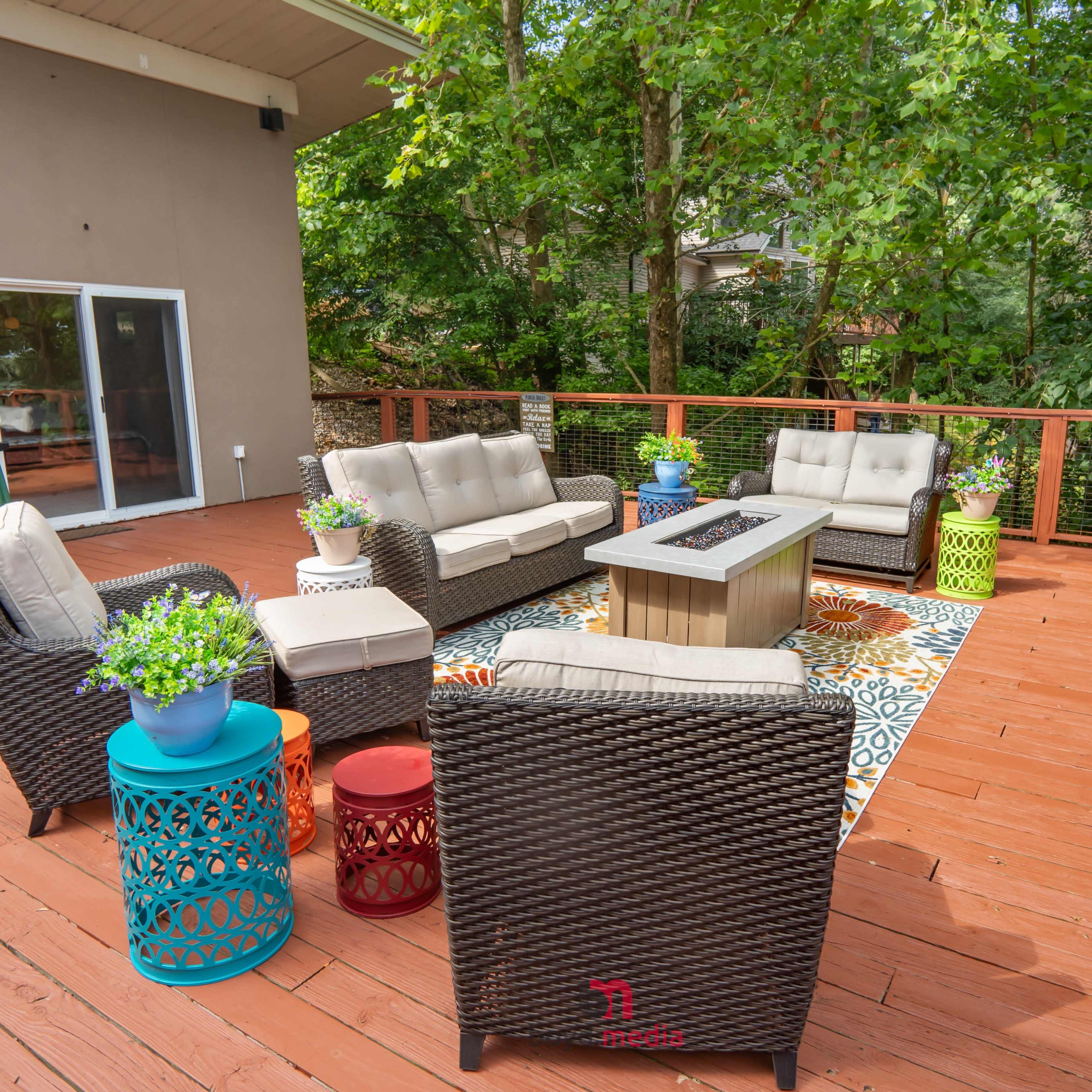 A furnished outdoor deck area with seating arrangements, a fire pit, and greenery in the background.