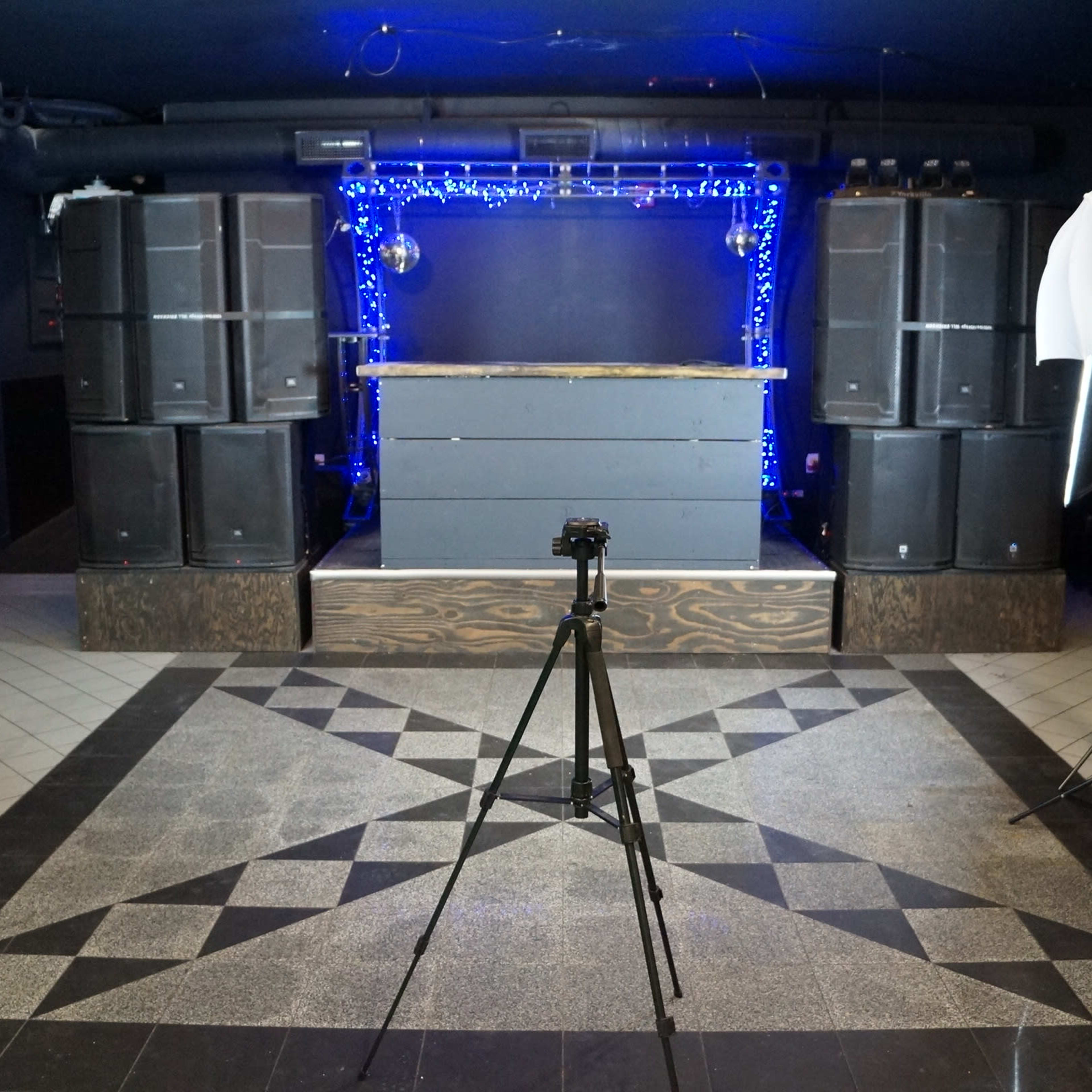 The scene features a photography setup in a dimly lit venue with a DJ booth, speakers, and lighting equipment arranged in front of a patterned floor.